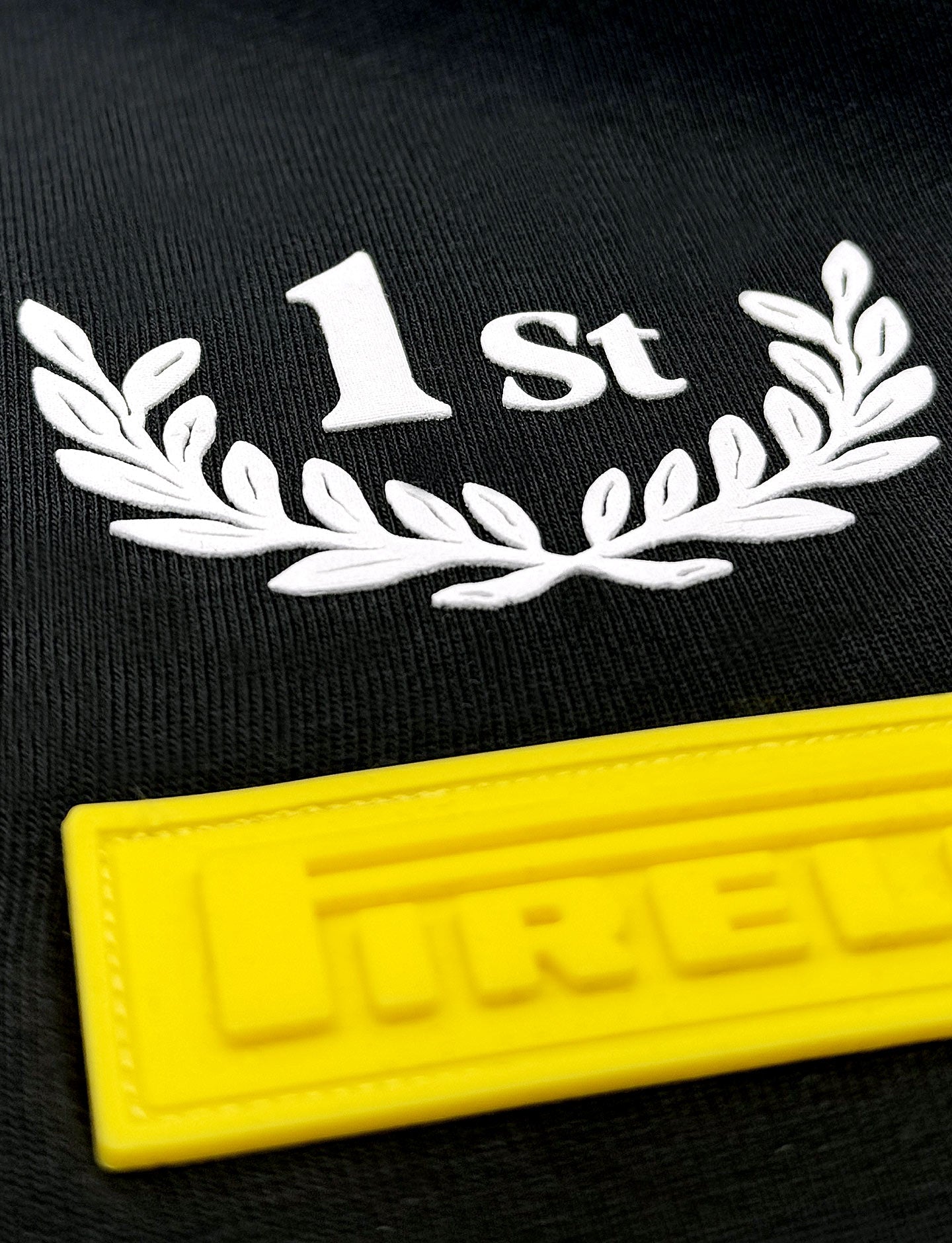 Black shirt with '1st' and laurel wreath design, featuring a yellow patch with 'Pirelli' branding.