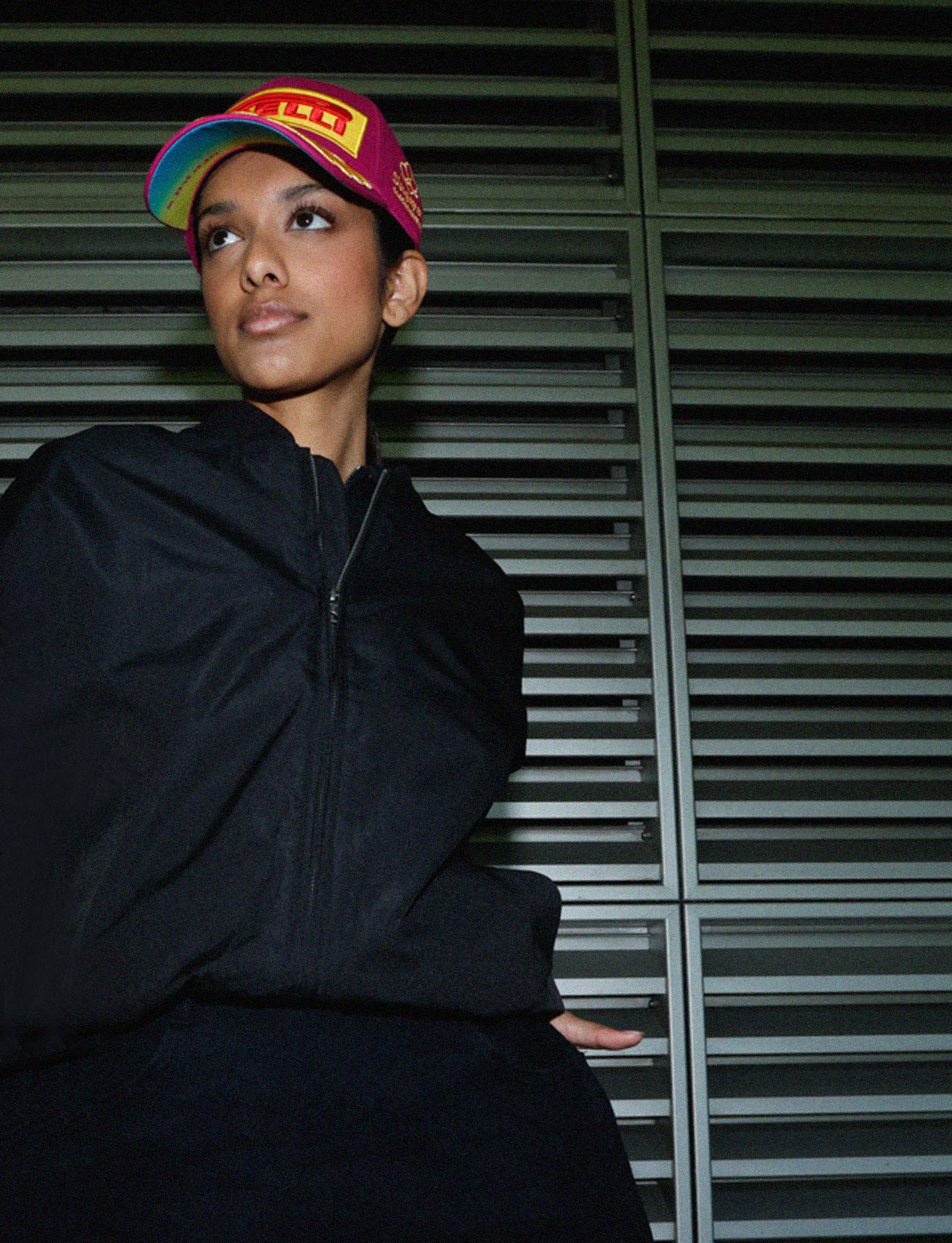 Person wearing a black jacket and Pirelli cap against a metallic shutter background