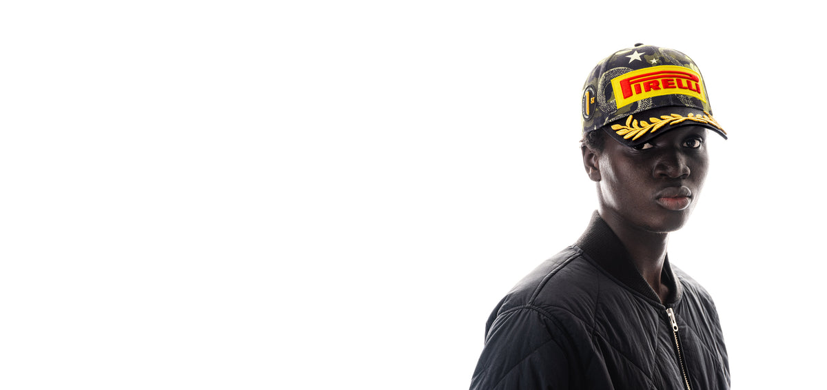 A man in a black jacket wearing a black Pirelli cap with yellow and red branding, photographed against a solid white background with him positioned on the right side.
