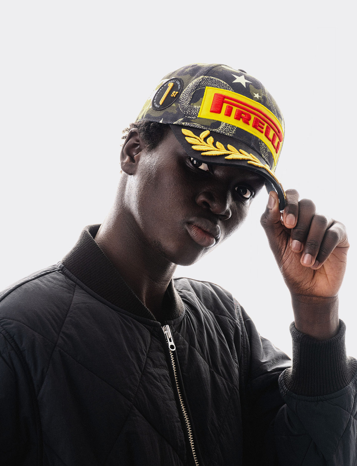 A man wearing a camouflage Pirelli F1® cap with yellow and red logo branding and a black quilted bomber jacket, posing against a light gray background.