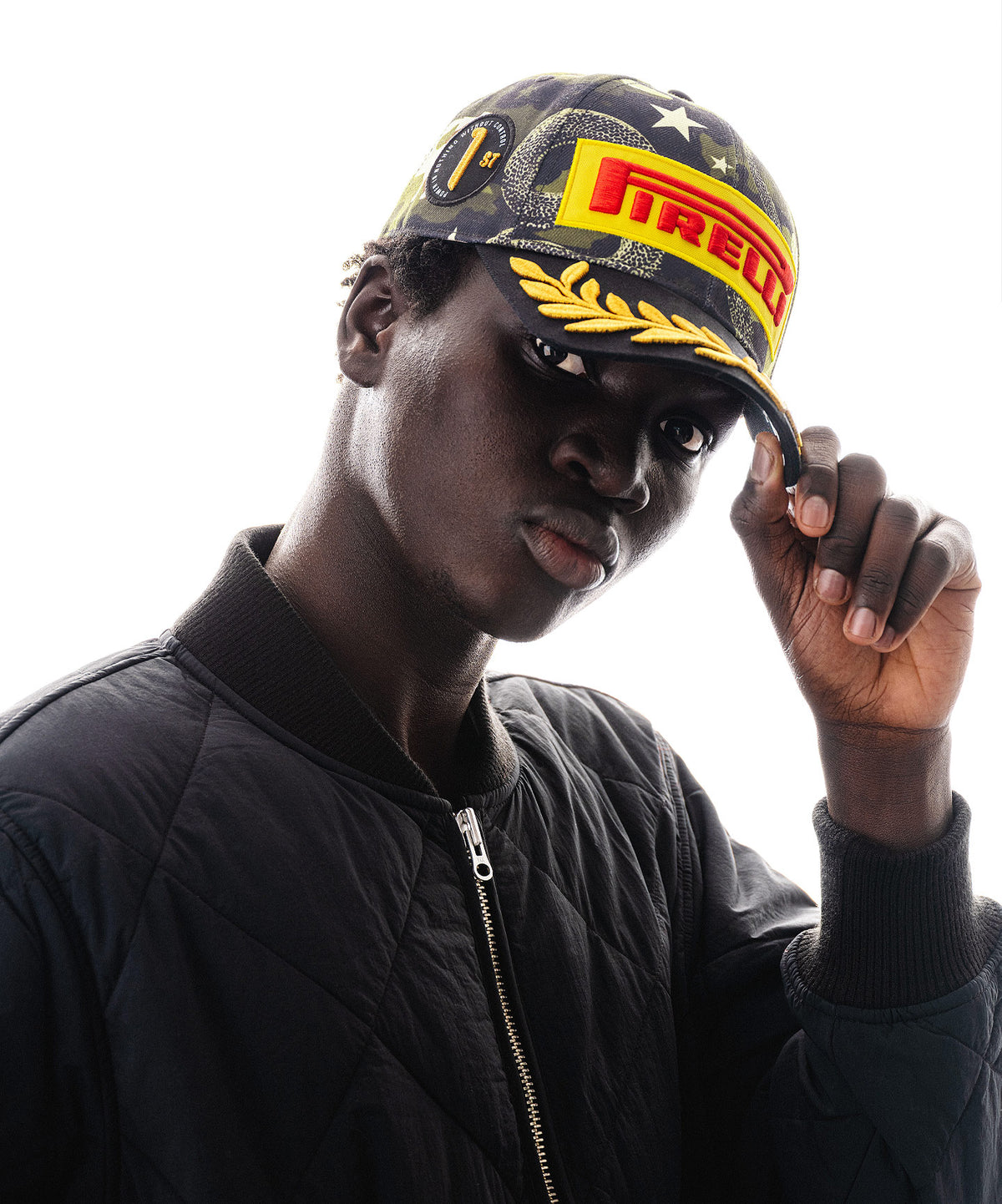 A man wearing a camouflage Pirelli F1® cap with yellow and red logo branding and a black quilted bomber jacket, posing against a light gray background.