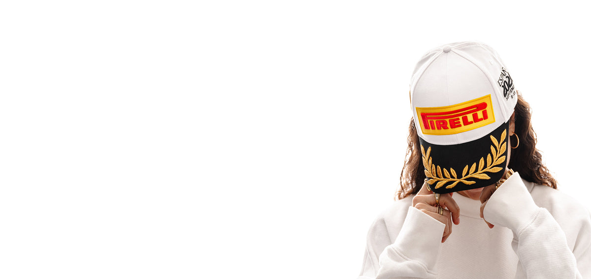 A person wearing a white Pirelli Podium Cap 2026 Madrid with a black and gold laurel band and a white hoodie, shown against a solid white background with negative space on the left.