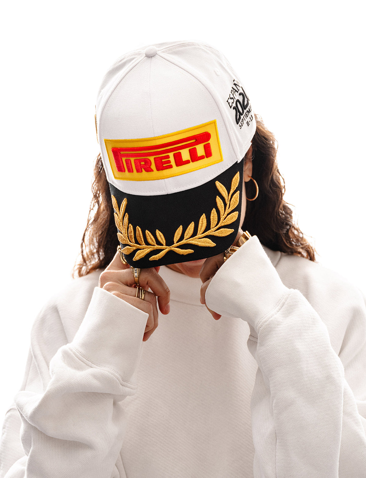 A person wearing a white Pirelli Podium Cap 2026 Madrid with a black lower band, gold laurel embroidery, and a red/yellow Pirelli logo patch, paired with a white sweatshirt against a plain white background.