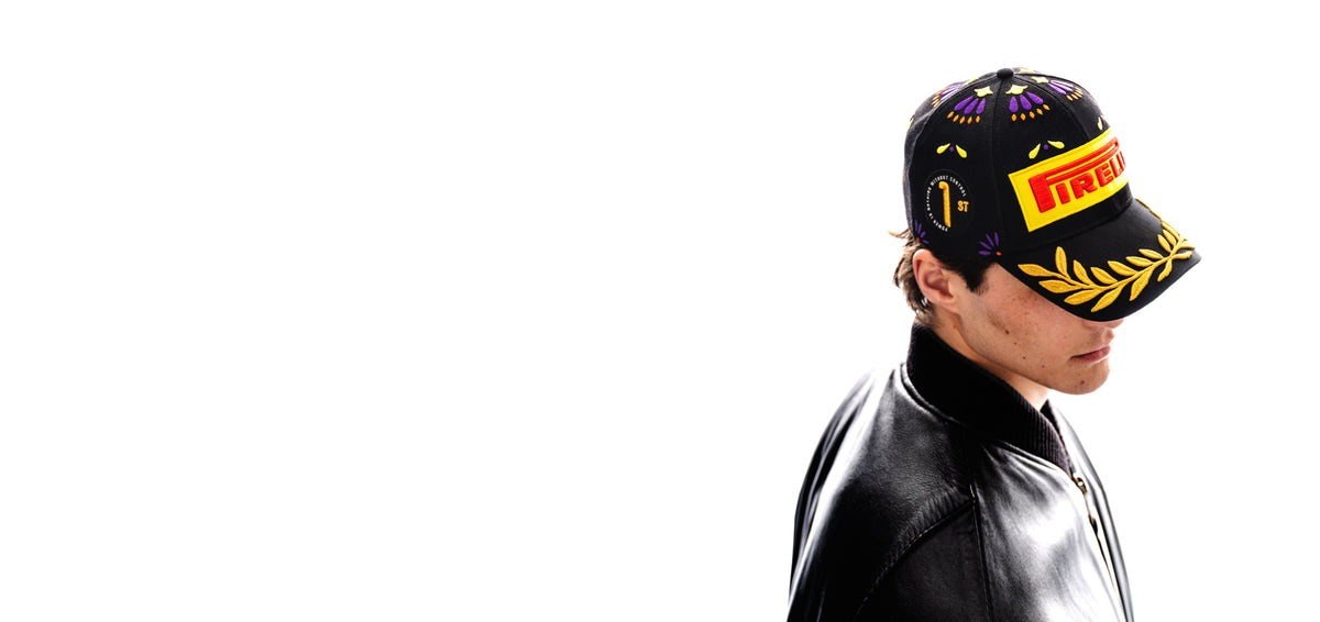 man wearing a black Pirelli Podium Cap 2026 Mexico, side view, with orange purple and yellow accents, a "1st" badge and laurels in gold embroidery.