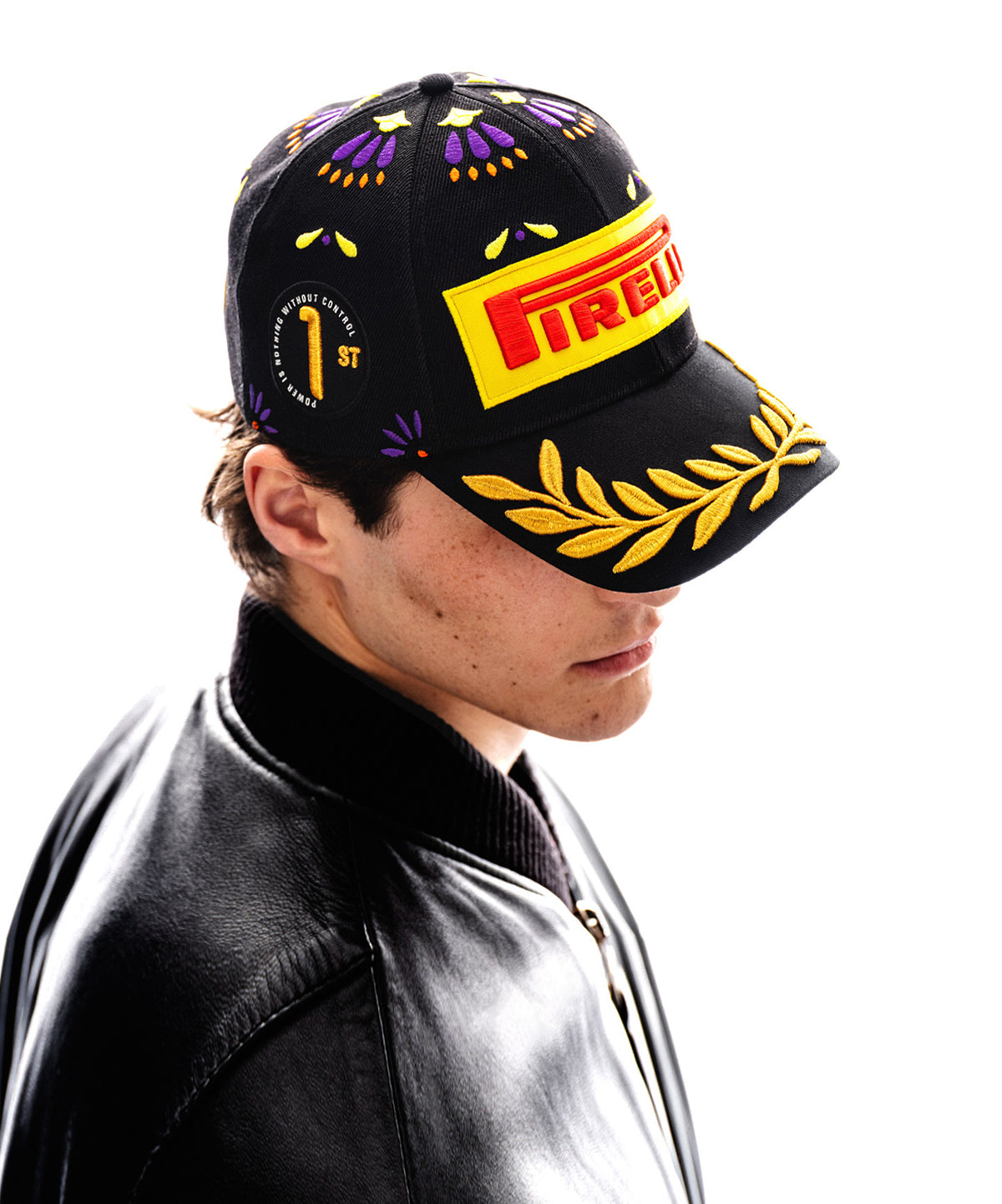 man wearing a black Pirelli Podium Cap 2026 Mexico, side view, with orange purple and yellow accents, a "1st" badge and laurels in gold embroidery.