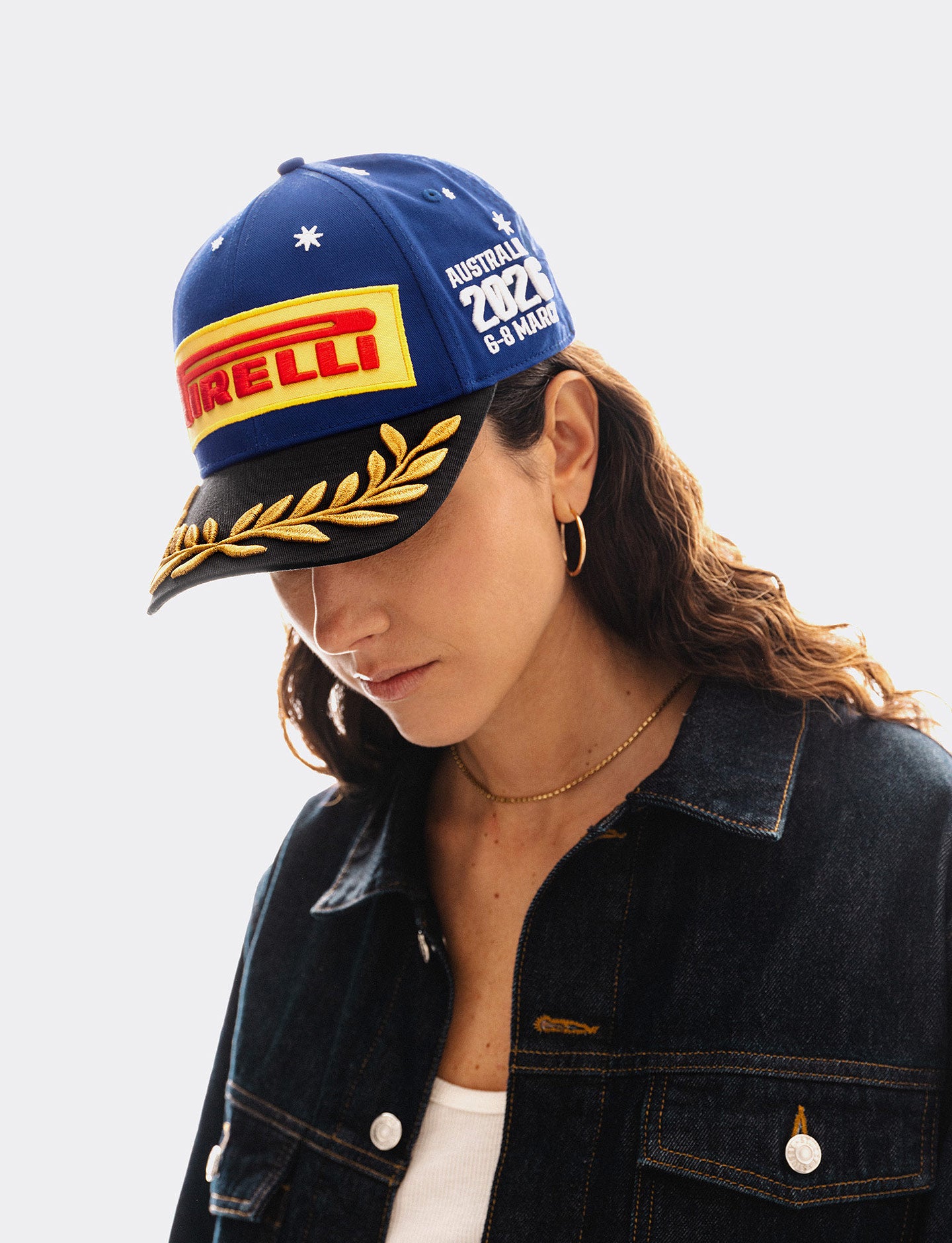 A woman wearing a blue Pirelli Podium Cap 2026 Australia, styled with a dark denim jacket over a white top, against a light gray studio background.