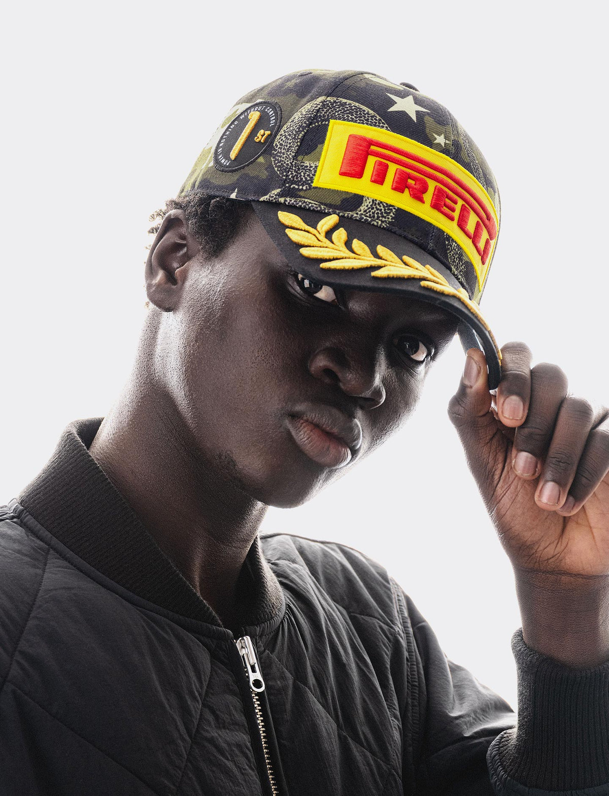 A close-up portrait of a man wearing a Pirelli Podium Cap 2026 Austin with yellow and red branding and gold laurel details, paired with a black quilted jacket against a light gray background.