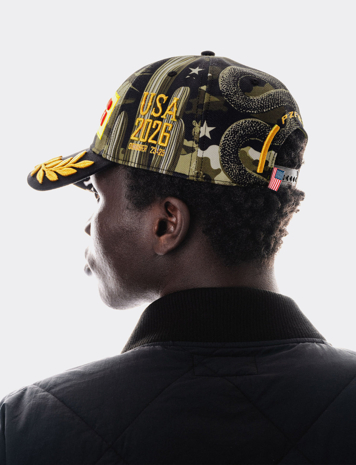 A man shown from the back and side wearing a Pirelli Podium Cap 2026 Austin with a complex graphic design. The cap features a camouflage pattern, a coiled rattlesnake, a gold eagle head, the text 'USA 2026', and a small American flag patch on the side.