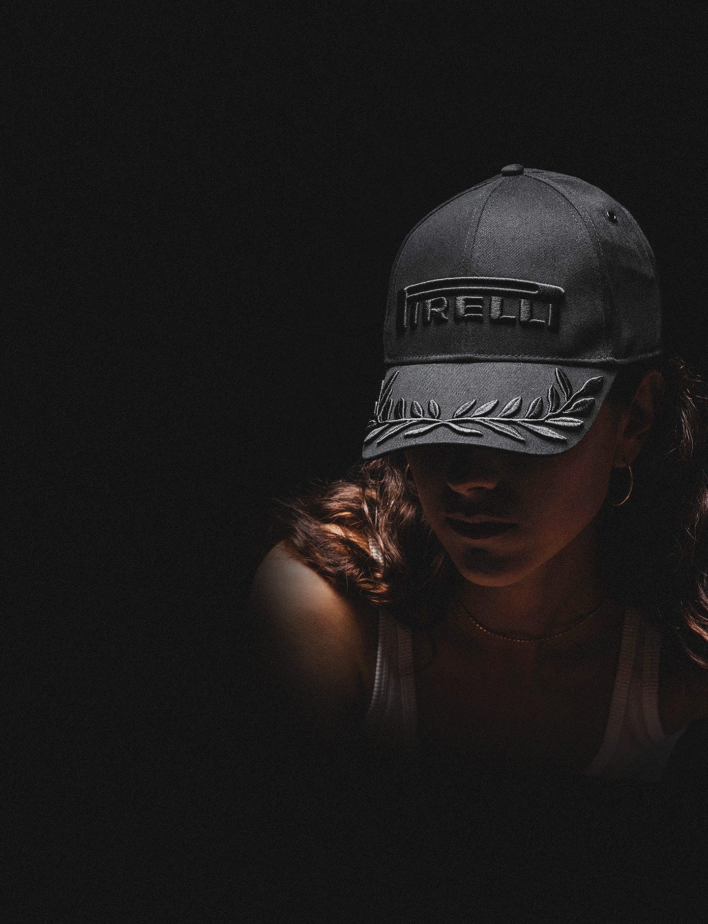A person wearing a black on black Pirelli baseball cap with a leaf embroidery on the brim, photographed in dramatic low light against a black background.