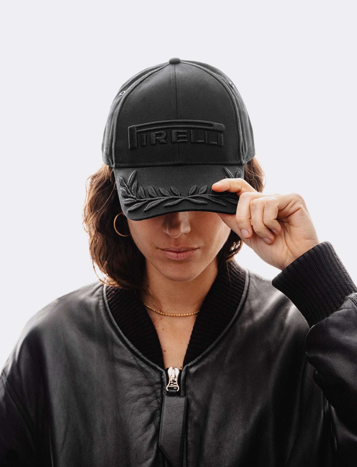 woman wearing a black on black Pirelli Podium Cap - front facing with her hand holding the visor