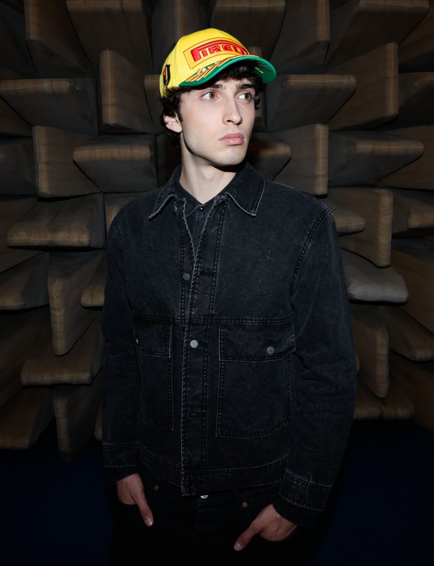 Person wearing a black jacket and yellow cap with a logo against a textured brown wall.