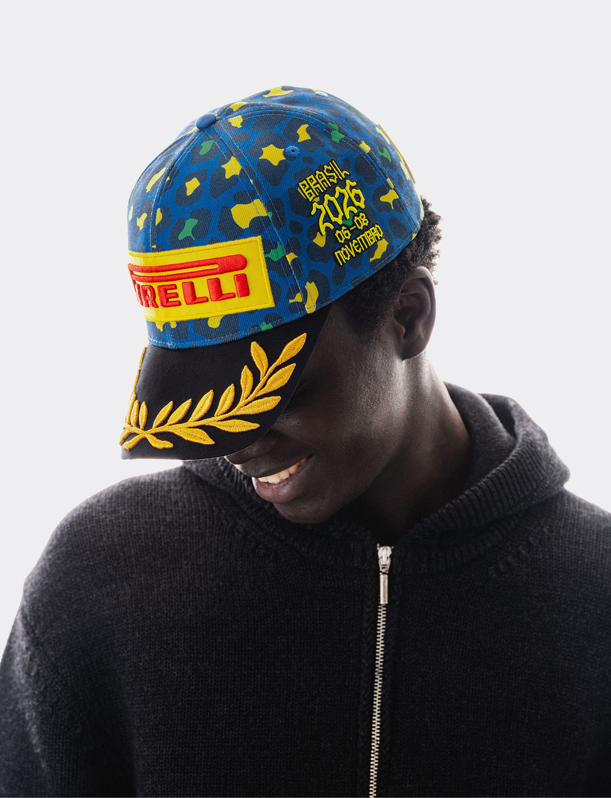 man wearing a Pirelli cap with blue, green and yellow camouflage on the crown, large Pirelli yellow and red logo on the front and golf laurel embroidery on the black brim.