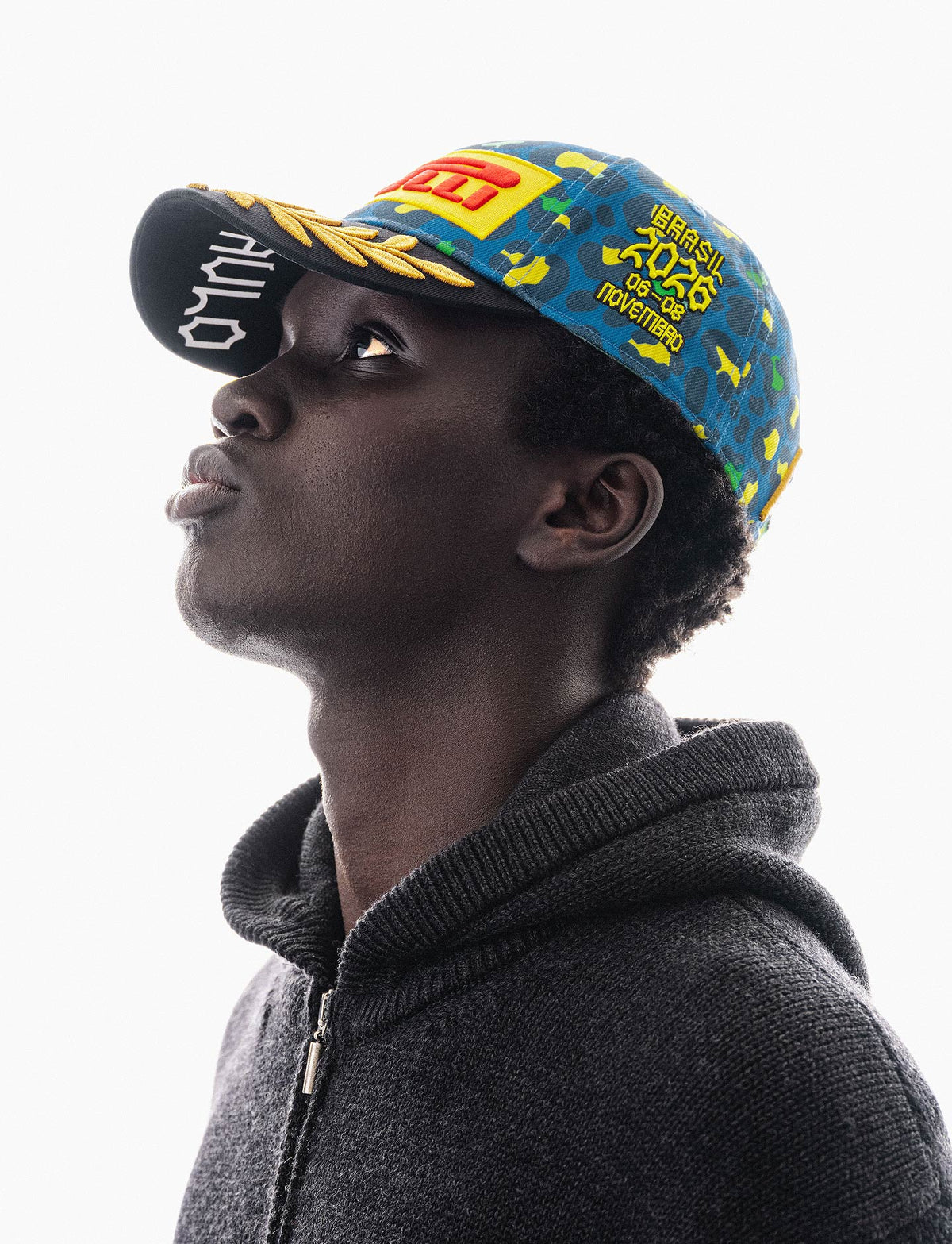 A man in profile wearing a dark gray zip-up hoodie and a colorful patterned Pirelli F1® baseball cap with yellow and blue and green jaguar spot graphics, set against a solid white background.