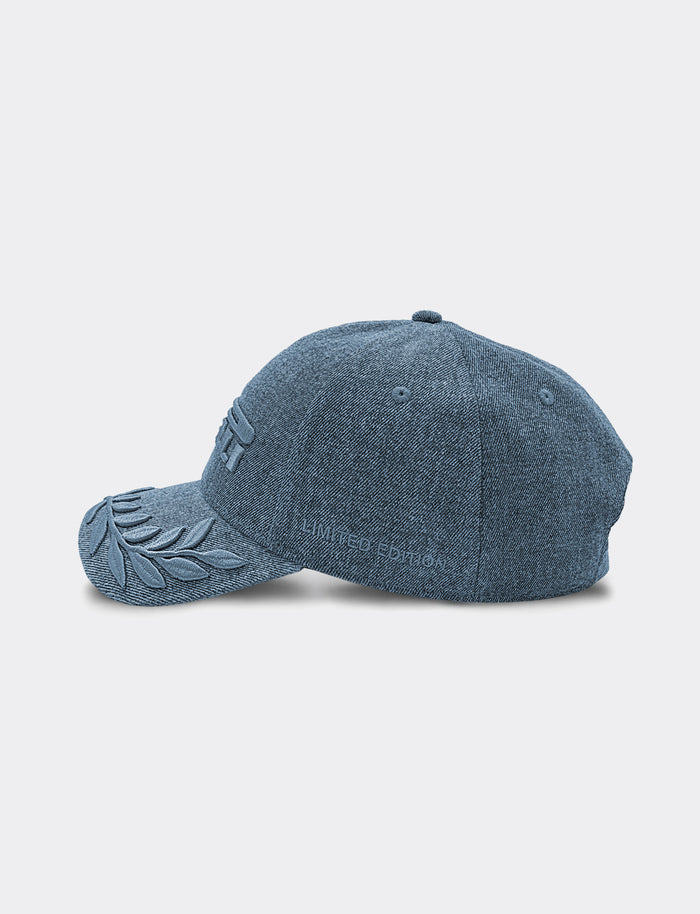 A blue denim Pirelli baseball cap shown from a side profile view against a light gray background. The cap features an embroidered laurel leaf design on the brim and subtle tonal embroidery "LIMITED EDITION" text on the side.