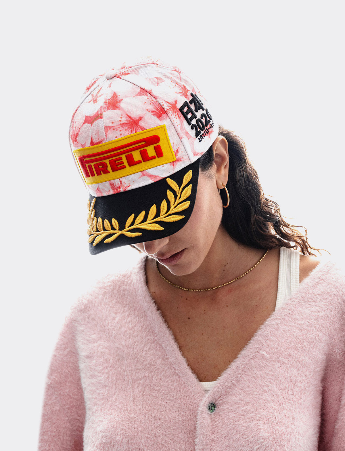 A woman wearing a pink fuzzy cardigan and a white and pink Pirelli Podium Cap 2026 Japan with a black brim with gold laurel embroidery, and a yellow and red Pirelli logo patch, photographed against a light gray background.