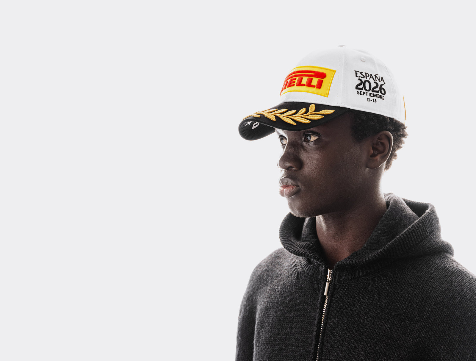 A man in a dark gray zip-up hoodie wearing a white Pirelli Podium Cap 2026 Madrid cap, shown in profile against a light gray background.