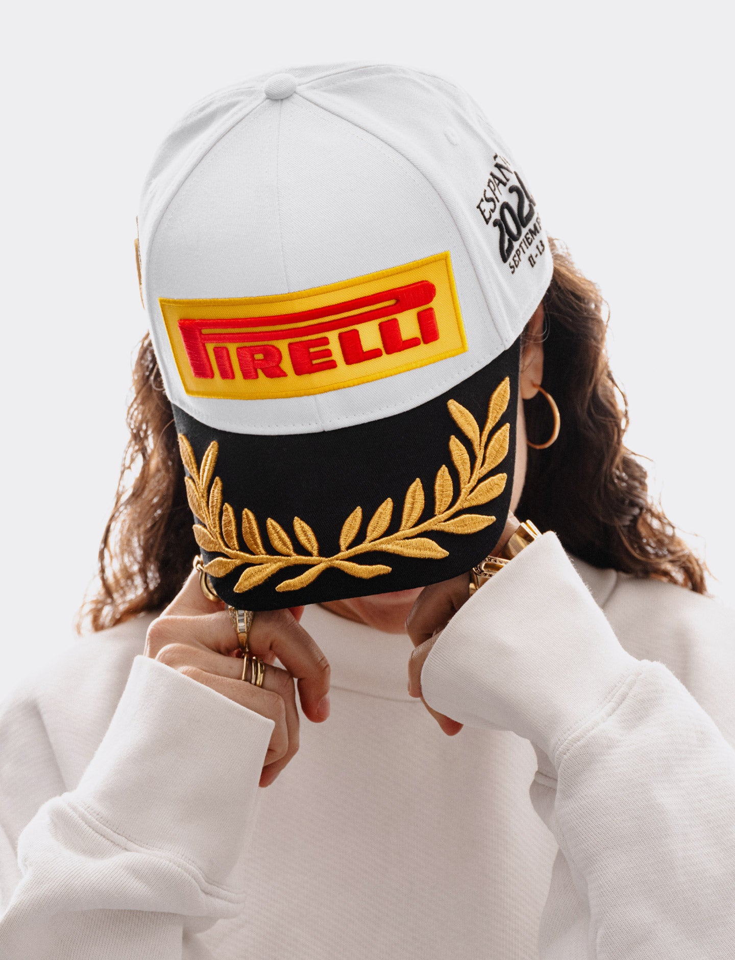 Woman wearing a Pirelli Podium Cap 2026 Madrid looking downwards and also wearing a white woven sweatshirt