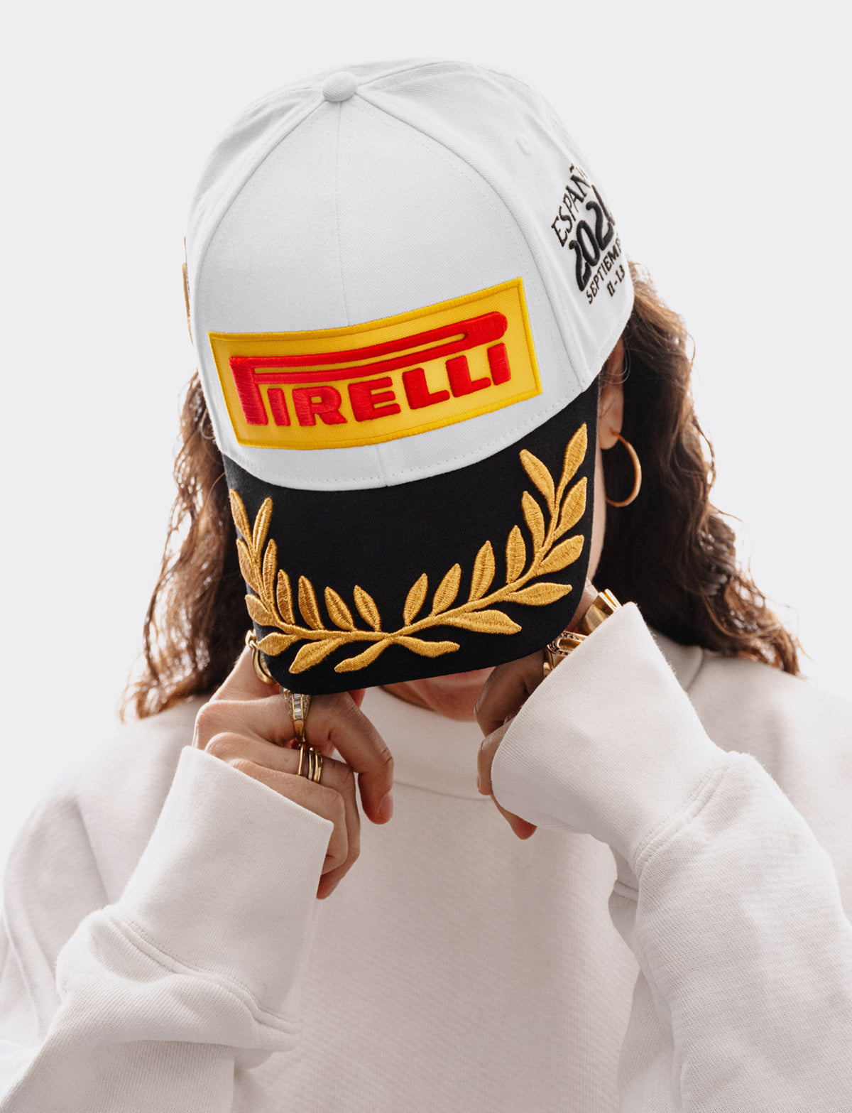 Woman wearing a Pirelli Podium Cap 2026 Madrid looking downwards and also wearing a white woven sweatshirt