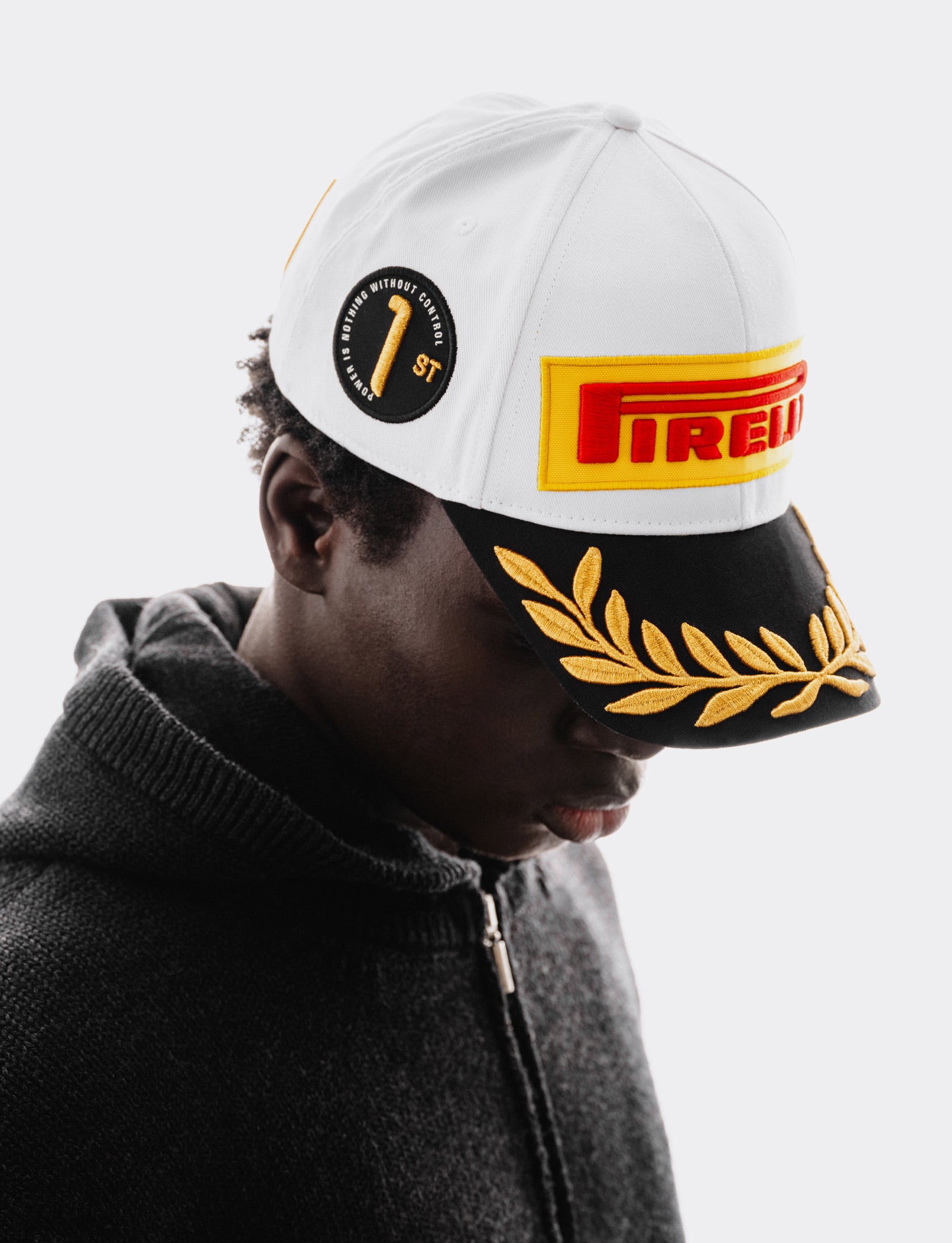 A person wearing a white Pirelli baseball cap with a black visor, gold laurel embroidery, and a black circular patch, paired with a dark gray zip-up hoodie against a light gray background.