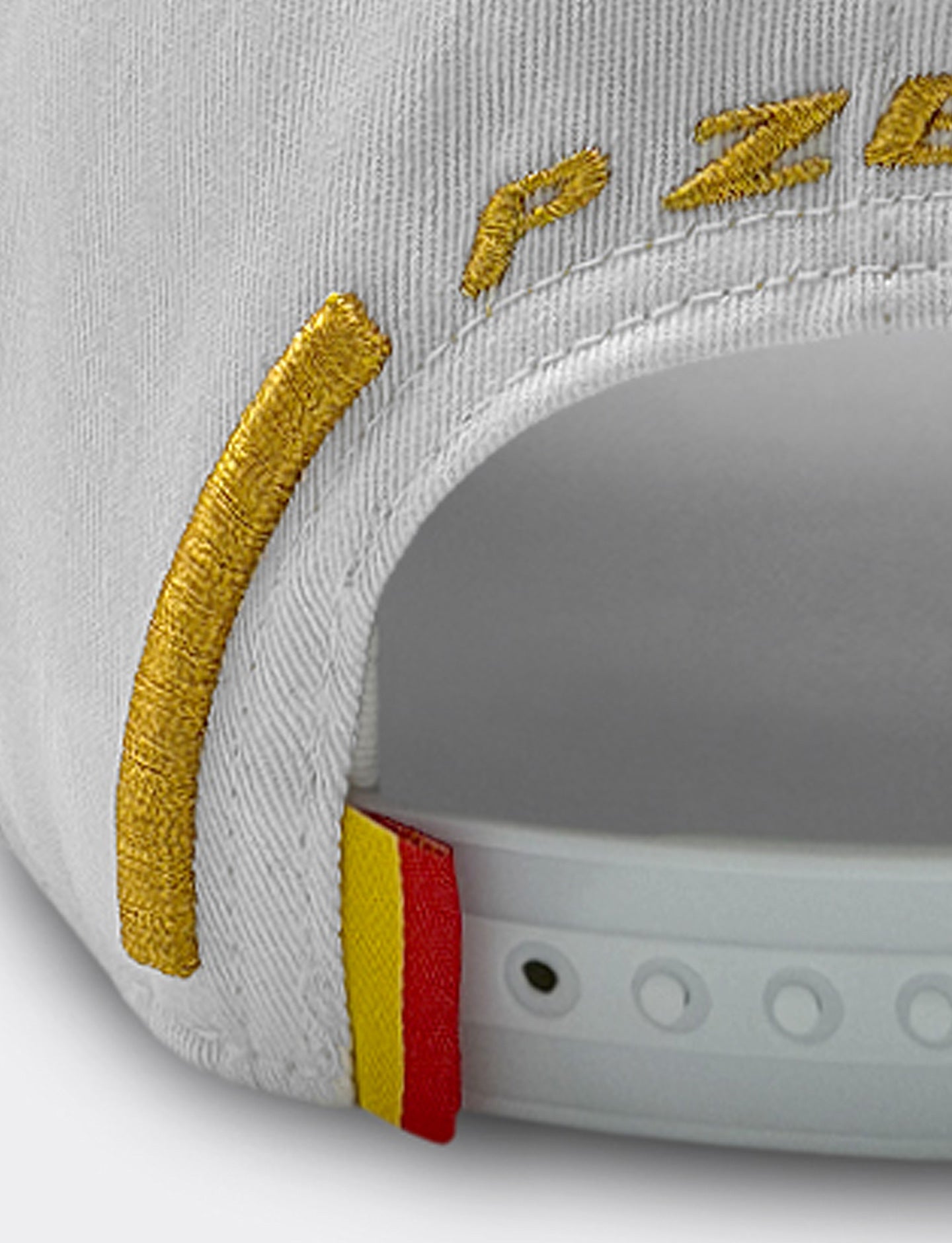 Close-up of a white baseball cap showing gold embroidered lettering and a small red and yellow Spanish flag on snapback closure.