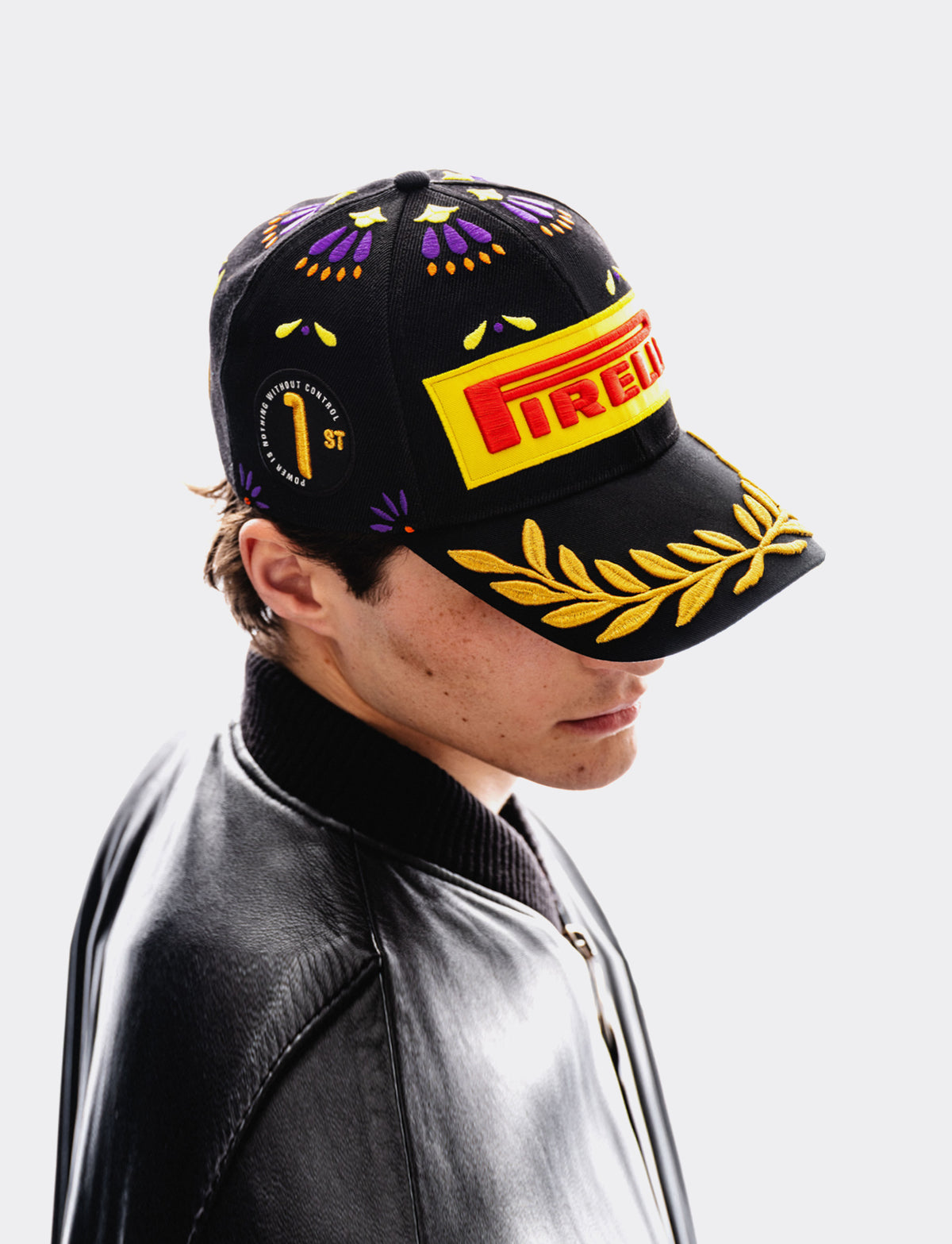 A black baseball cap with purple and yellow embroidered patterns, a yellow Pirelli logo patch on the brim, and a gold laurel leaf embroidery, worn by a model in a black leather jacket against a light gray background.