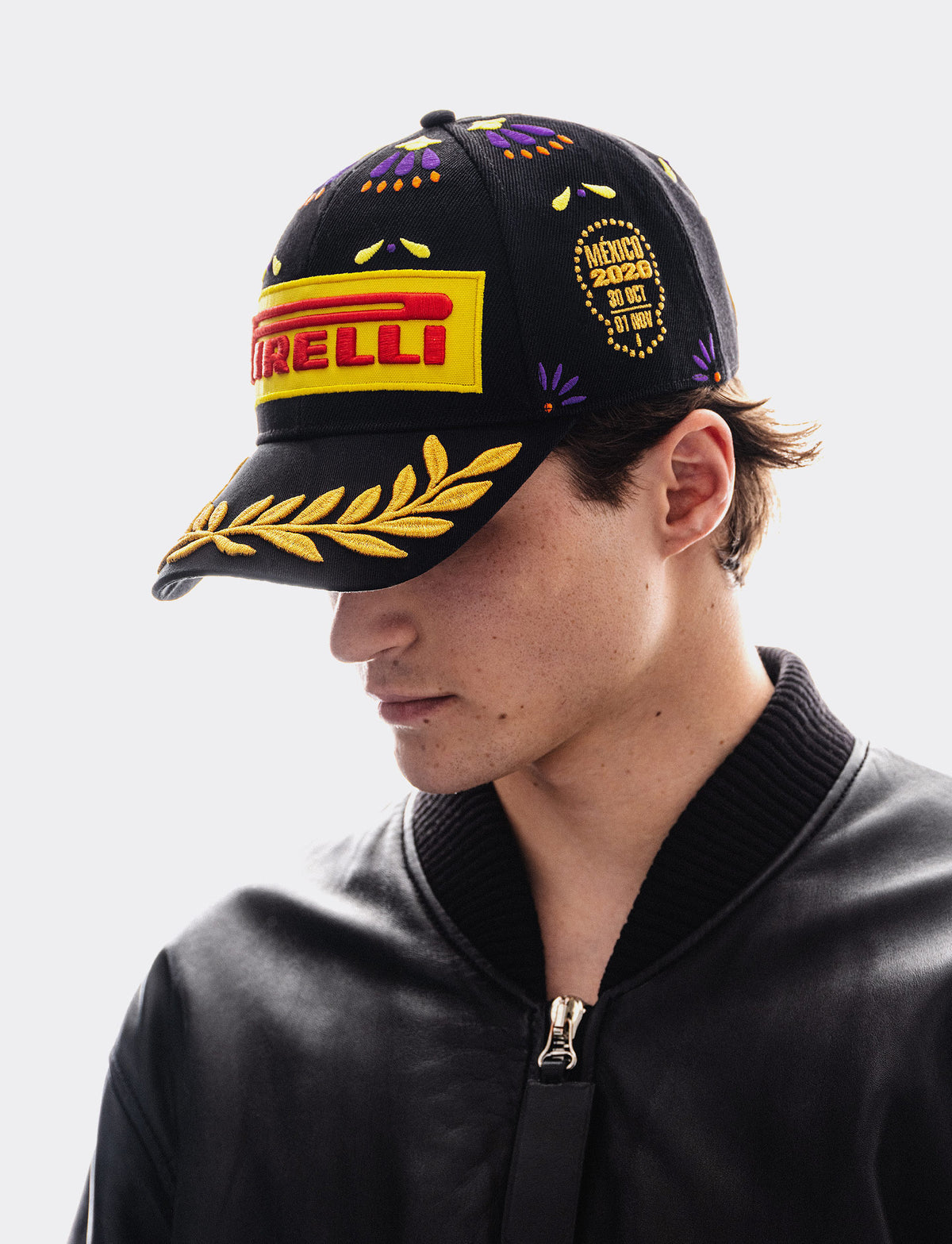 Man wearing a black Pirelli Podium Cap 2026 Mexico baseball cap featuring a prominent yellow and red embroidered logo on the front, a gold laurel leaf embroidery on the brim, and multicolor geometric patterns on the crown.