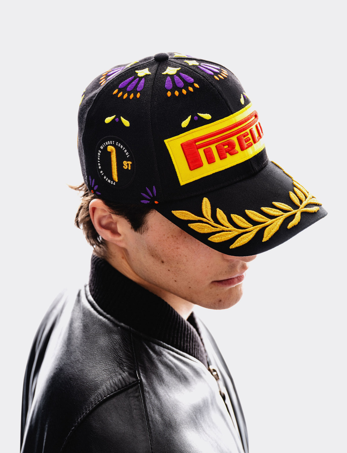 A man in a black leather jacket wearing a black Pirelli baseball cap with yellow and purple embroidered details, a yellow Pirelli logo patch on the brim, and a gold laurel wreath embroidery on the brim.