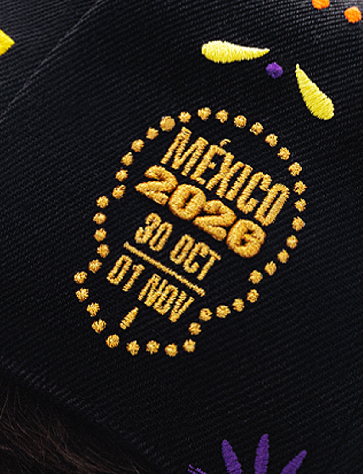 Close-up of a black knit fabric featuring a circular gold embroidered design with the text 'MEXICO 2026 30 OCT 01 NOV' and small yellow and purple embroidered accents.