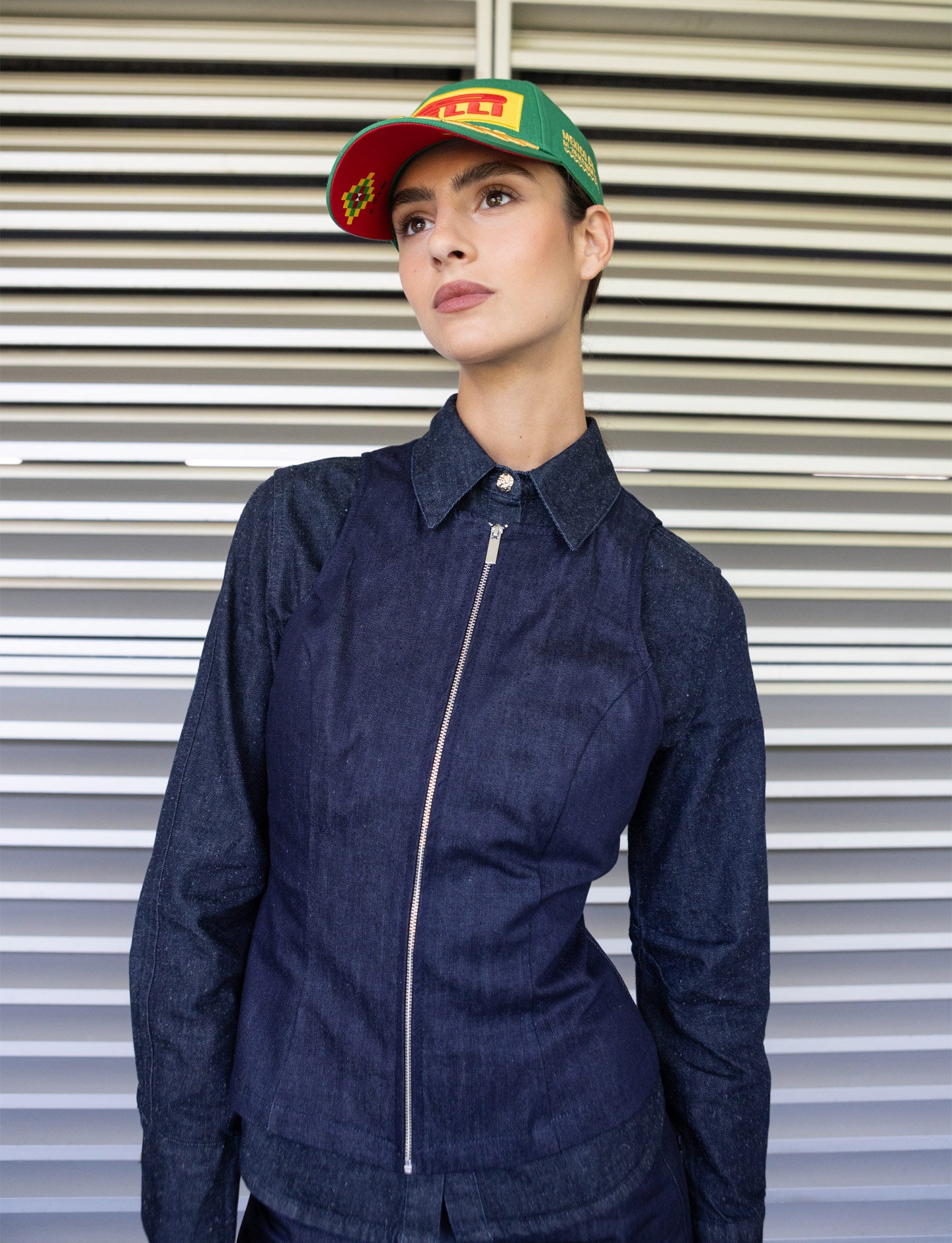 Person wearing a navy jacket and green Pirelli Mexico cap against a metallic shutter background