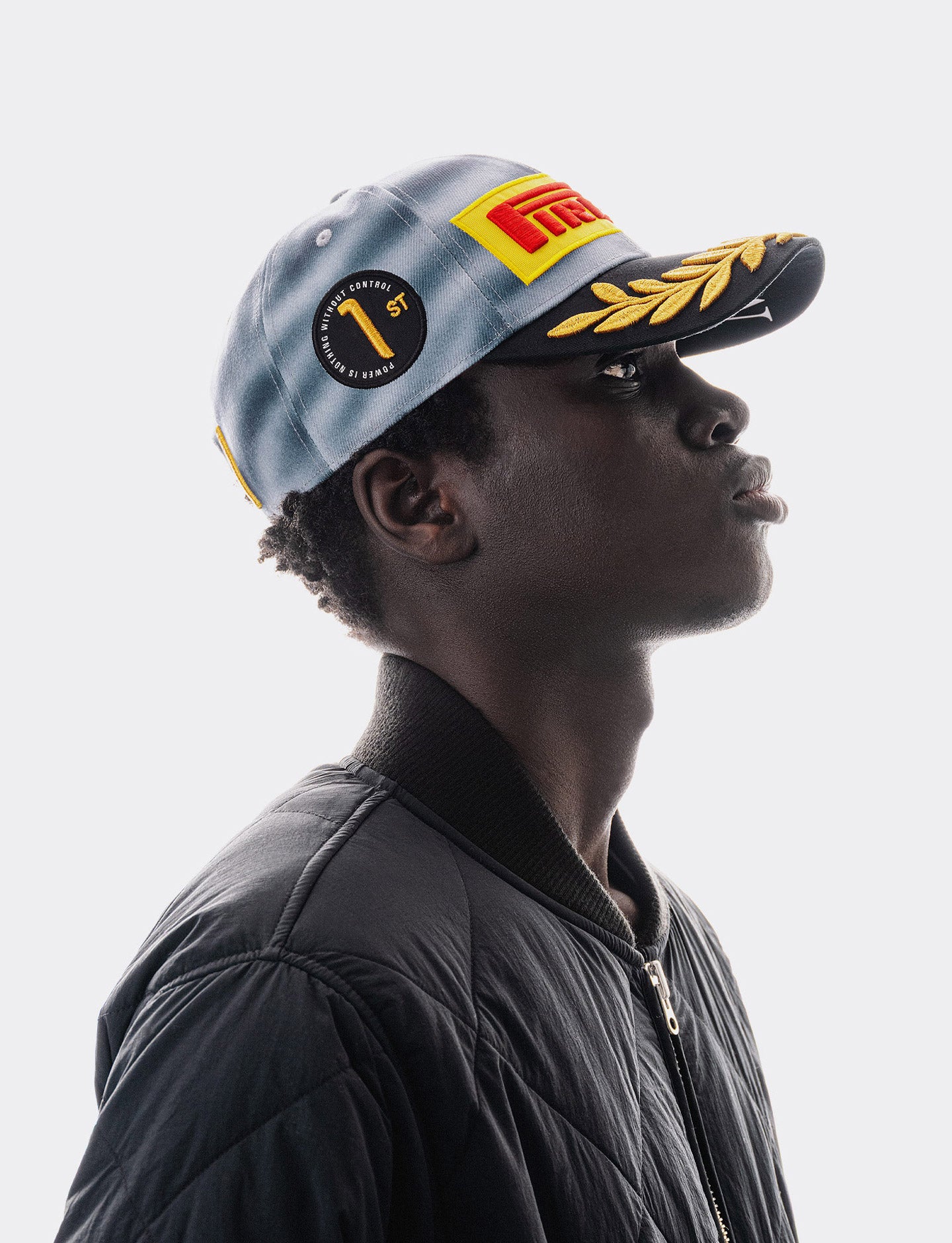 A man in profile wearing a light blue Pirelli Podium Cap 2026 Miami with a yellow and red Pirelli logo patch and gold laurel embroidery on the brim, paired with a black quilted jacket against a light gray background.
