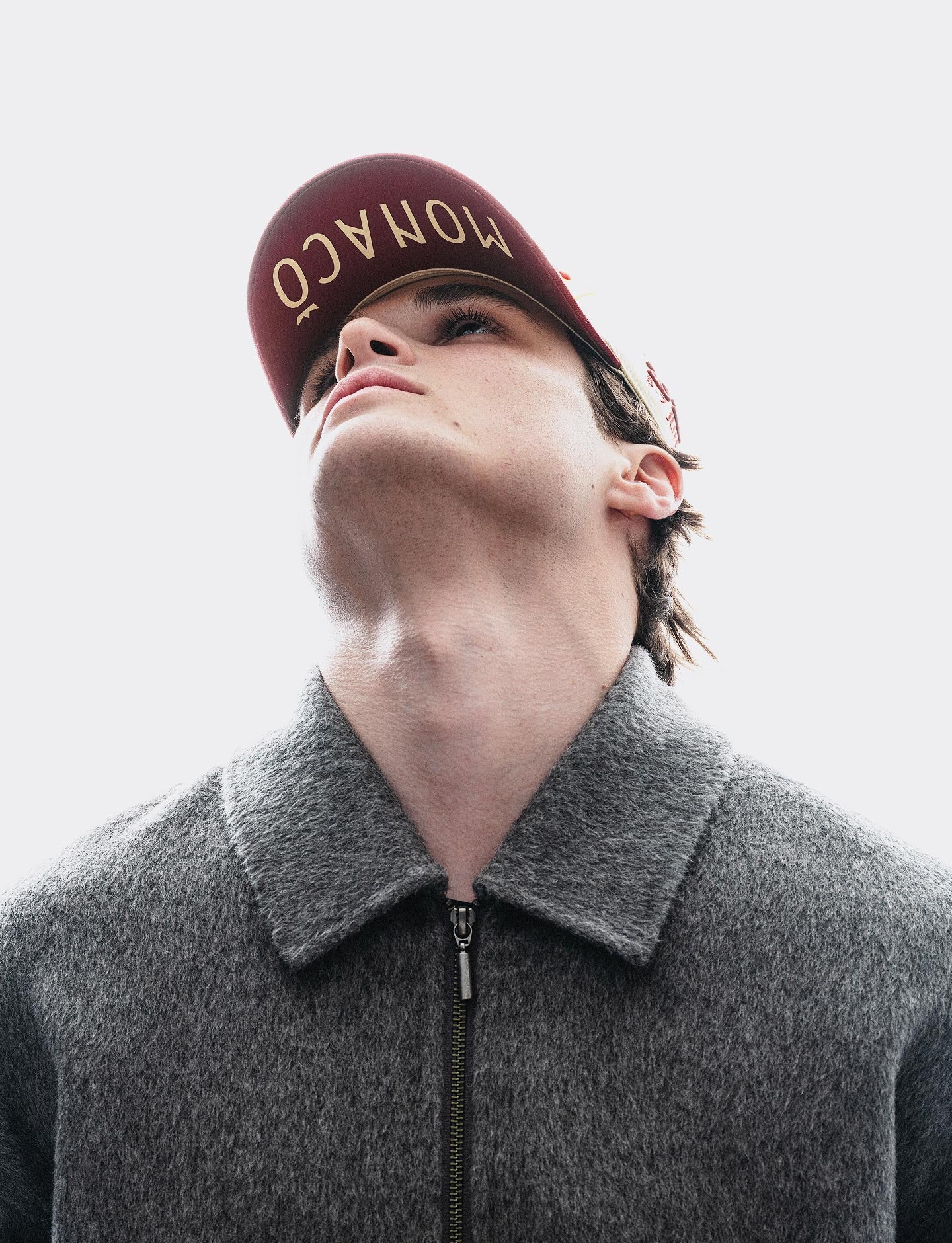 A man wearing a Pirelli Podium Cap 2026 Monaco with 'MONACO' printed in gold on the front, looking upward against a bright white sky. He is wearing a textured gray zip-up jacket.