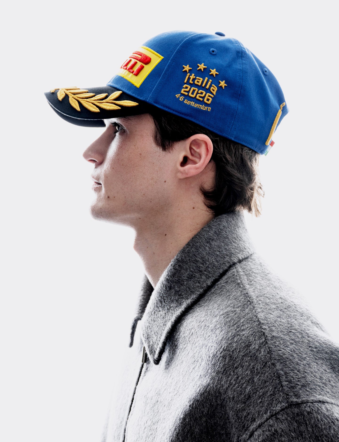 A man in profile wearing a blue Pirelli Monza 2026 podium cap featuring 'F1 Italia 2026', paired with a gray textured jacket against a light gray background.