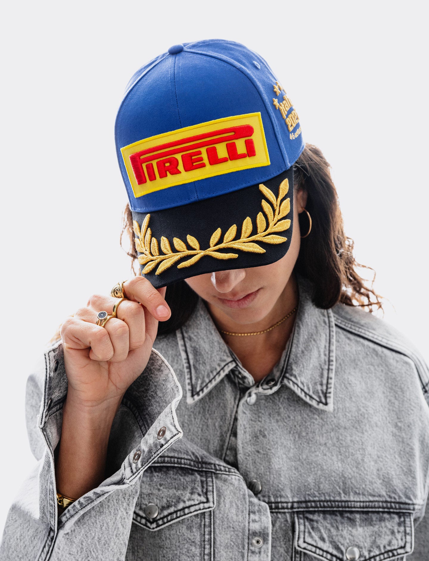 A person wearing a blue and black Pirelli Monza 2026 cap with gold embroidery, partially covering their face, paired with a gray denim jacket against a light gray background.