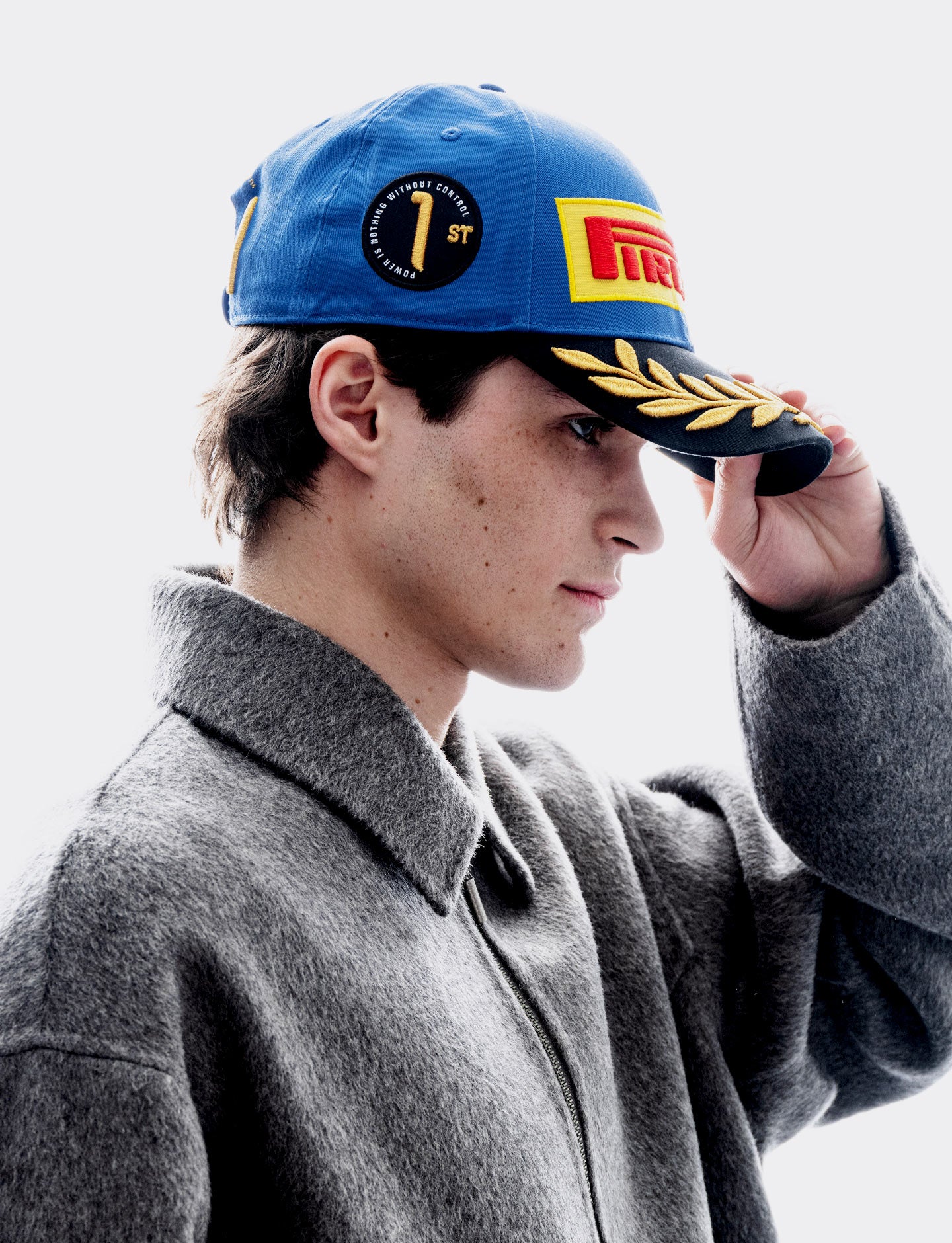 A man in a gray coat wearing a blue Pirelli Monza 2026 cap tipping the brim with his hand against a light gray background.