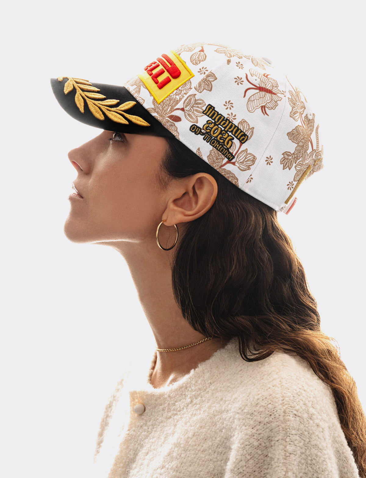 A woman in profile wearing a white Pirelli Podium Cap 2026 Singapore baseball cap with burgundy floral patterns, a black brim with gold laurel embroidery, and yellow/red Pirelli logo patch on the front, set against a solid white background.