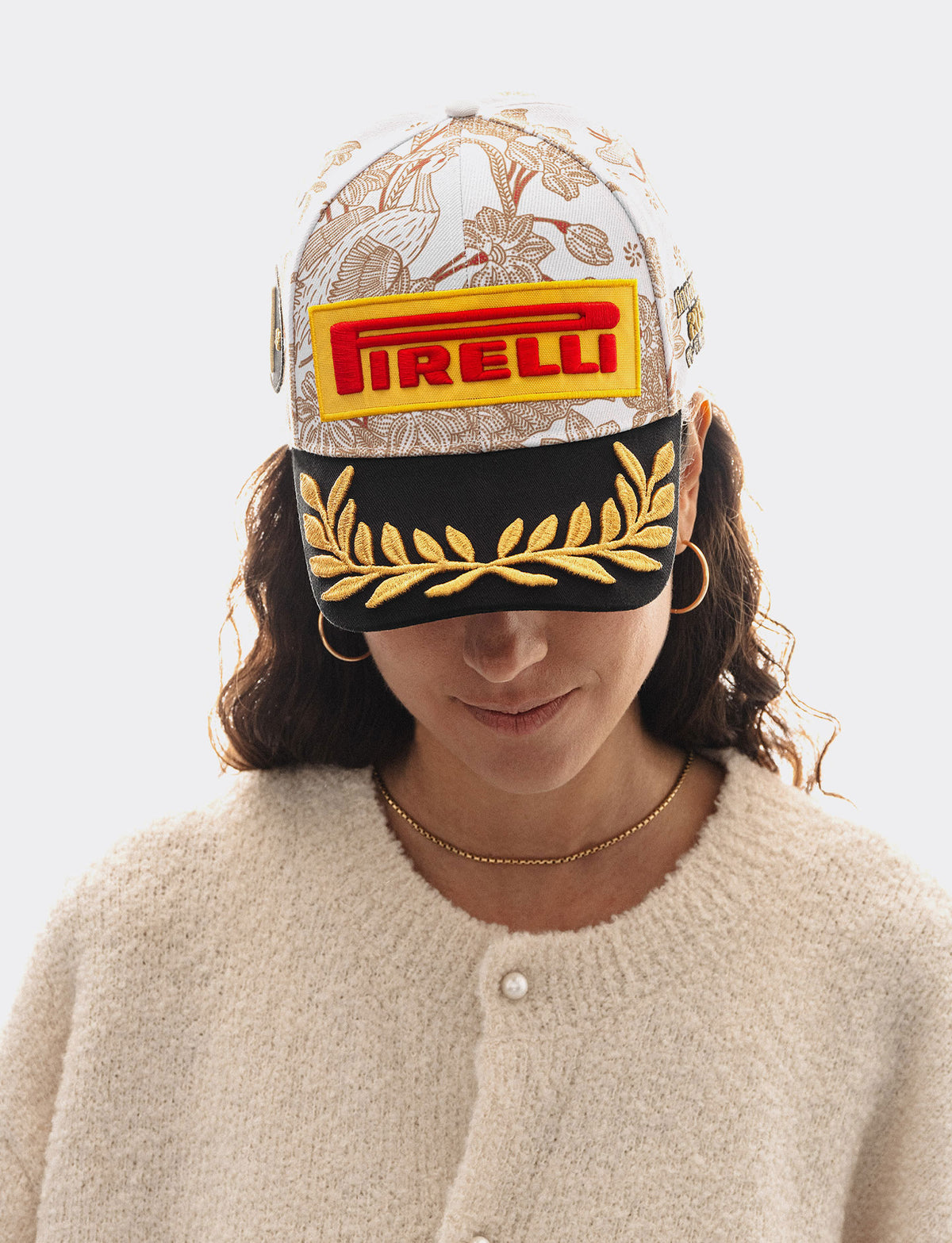 A woman wearing a white Pirelli Podium Cap 2026 Singapore baseball cap with burgundy floral patterns, a black brim with gold laurel embroidery, and yellow/red Pirelli logo patch on the front, set against a solid white background.