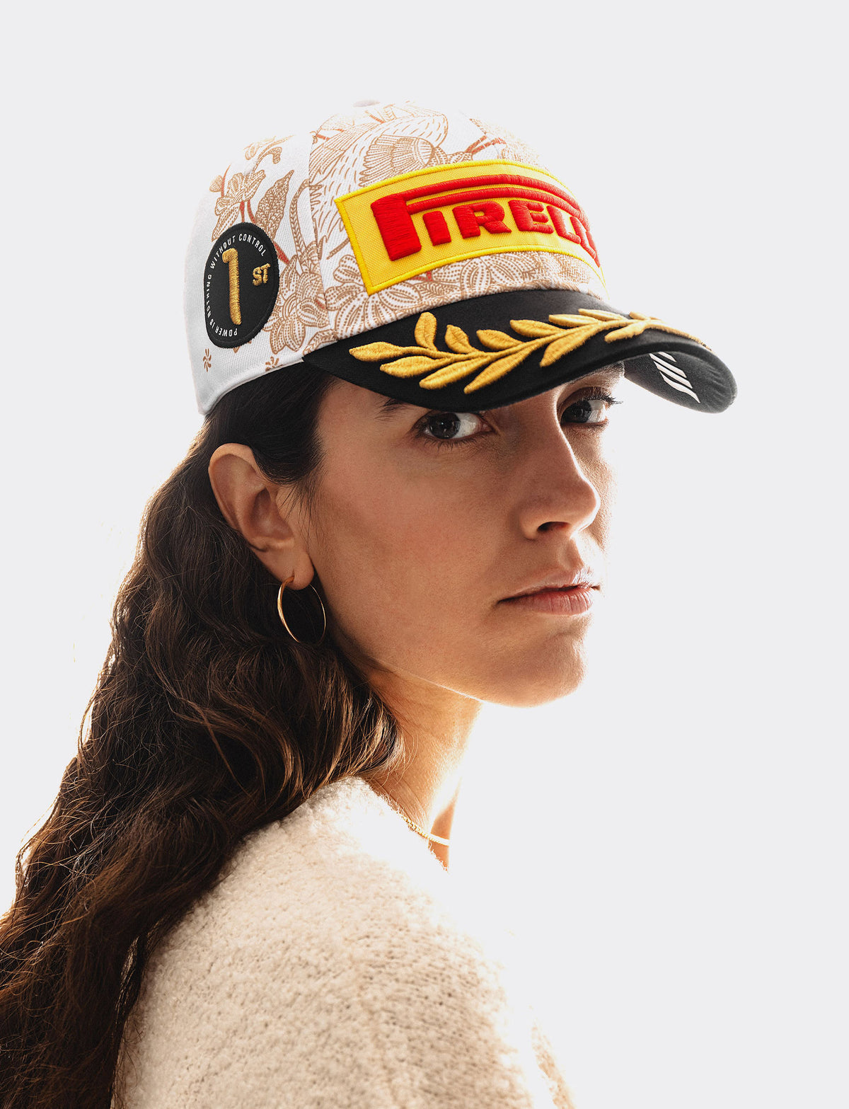 A woman wearing a Pirelli Podium Cap 2026 Singapore with a white floral pattern, a red and yellow Pirelli logo patch on the front, and a black brim with gold embroidered laurel leaves, shown against a bright white background.