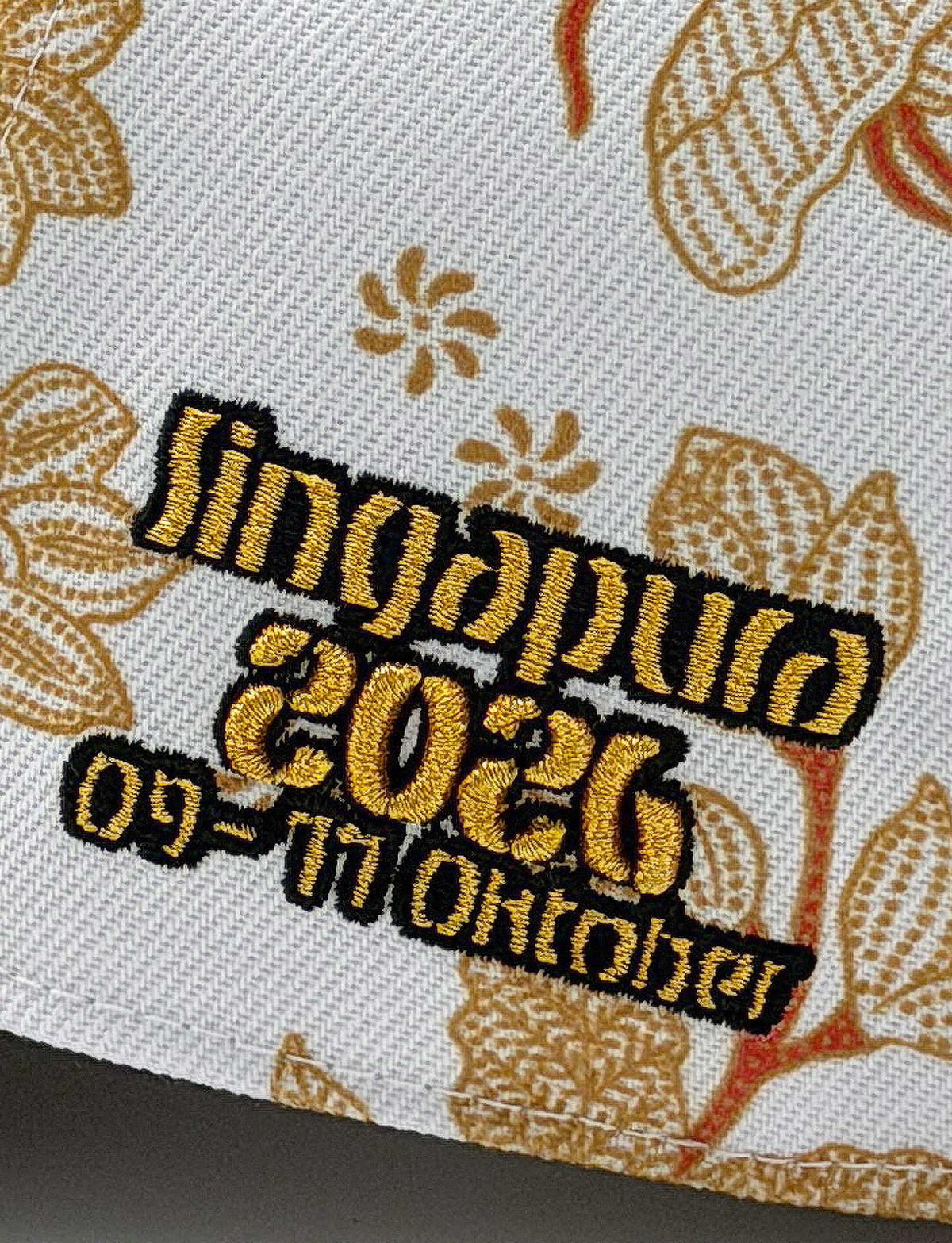 Closeup of Pirelli Podium Cap 2026 Singapore showing gold embroidered "Singapura 2026 09-11 Oktober" on a white cap with orange/brown floral print.
