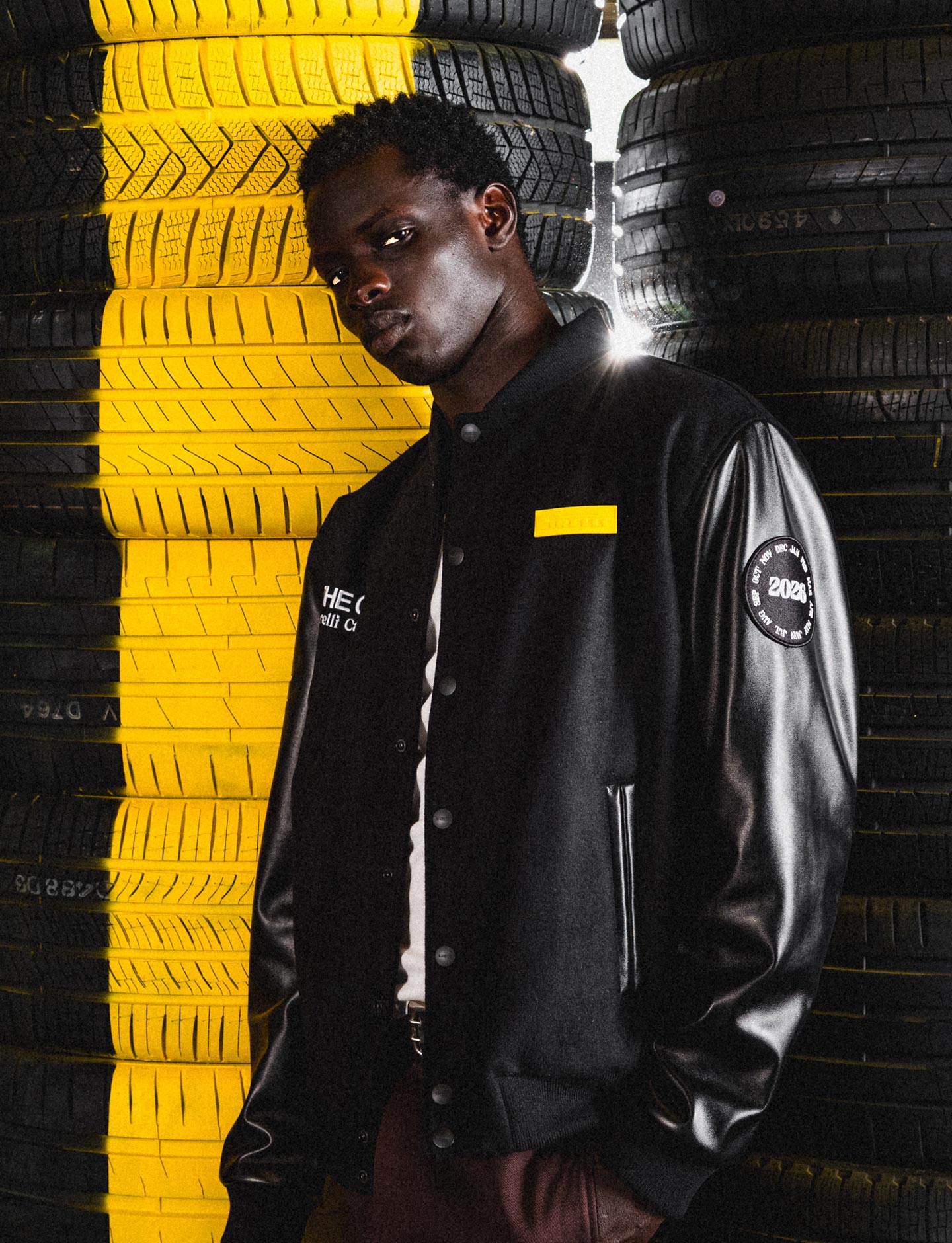 Person wearing a black Pirelli The Cal jacket with logos standing against a background of stacked tires, some yellow.