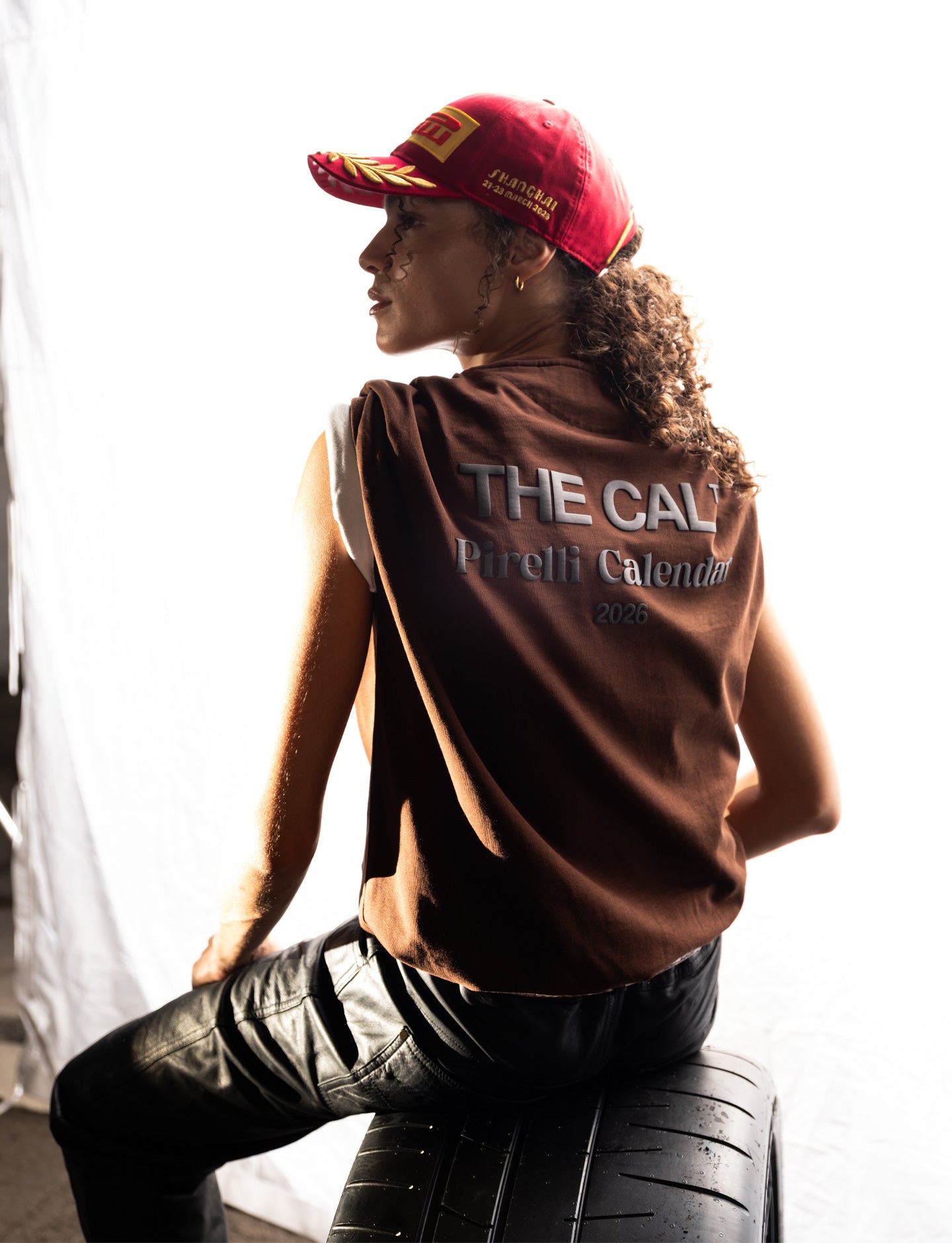 Person wearing a brown Pirelli tee with 'The Cal' text, sitting on a tire against a white background