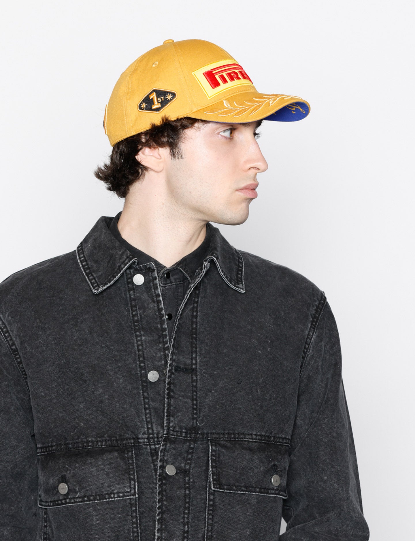 Person wearing a gold Pirelli cap with a logo and a black denim jacket on a white background