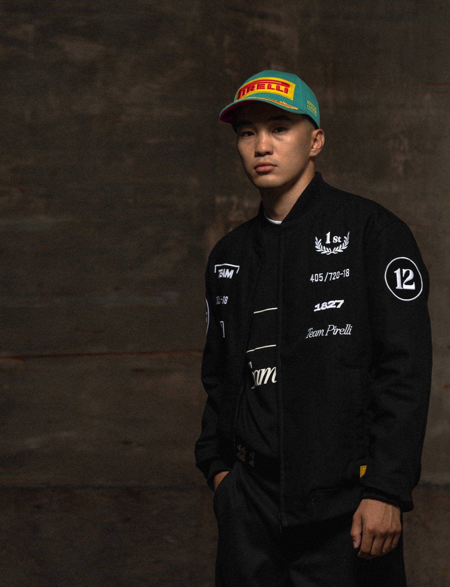 Person wearing a Pirelli racing jacket and cap against a brown stone background