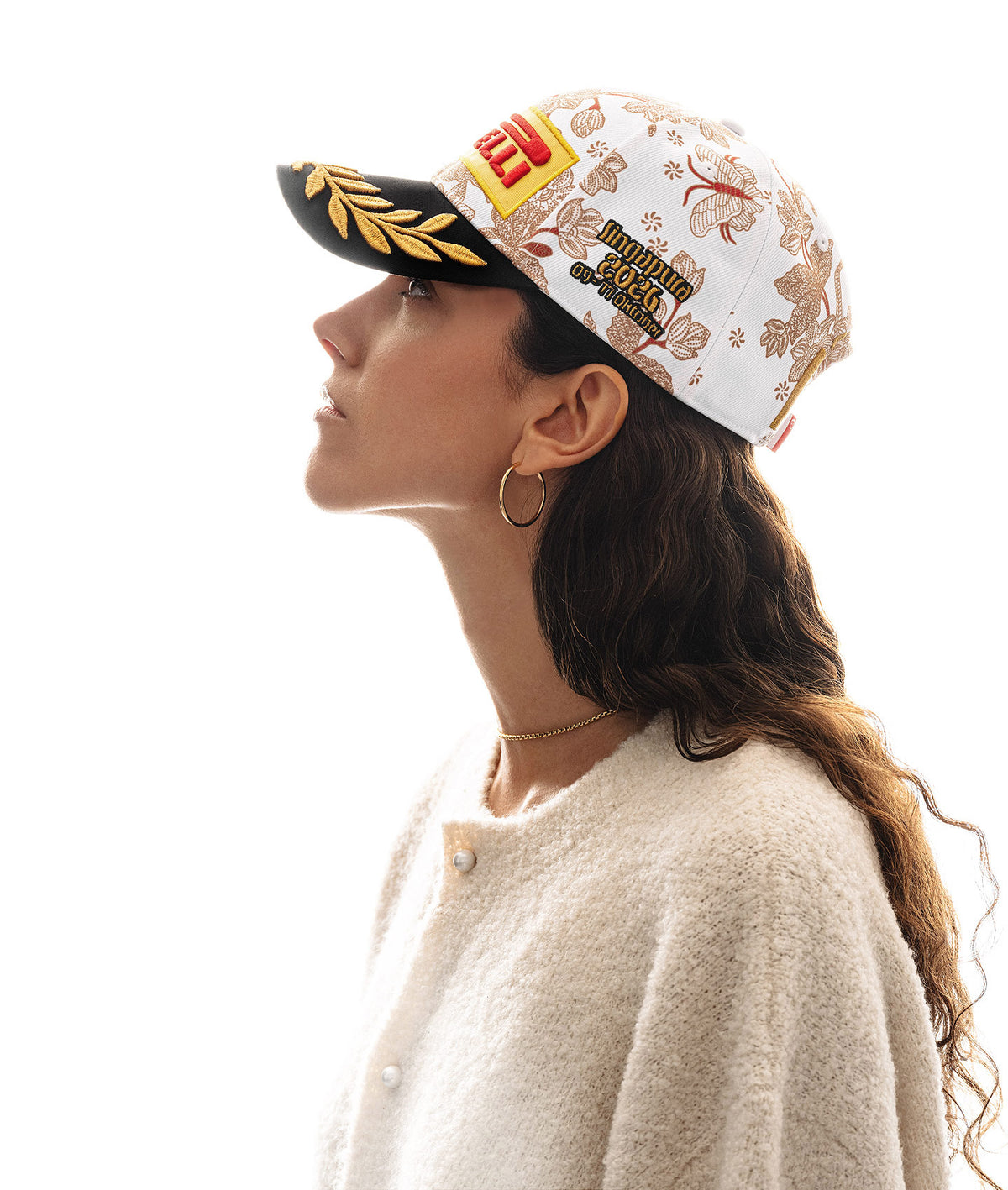 Side profile of a curly haired woman looking to upper left wearing a Pirelli Podium Cap 2026 Singapore with orange/brown floral pattern on white crown and with a black brim with gold laurel embroidery.