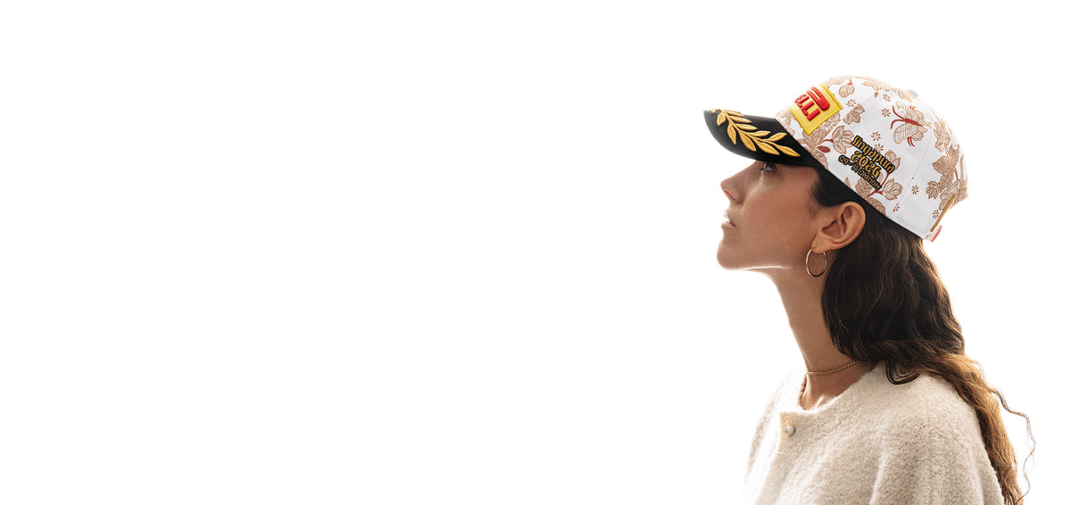 Side profile of a curly haired woman looking to upper left wearing a Pirelli Podium Cap 2026 Singapore with orange/brown floral pattern on white crown and with a black brim with gold laurel embroidery