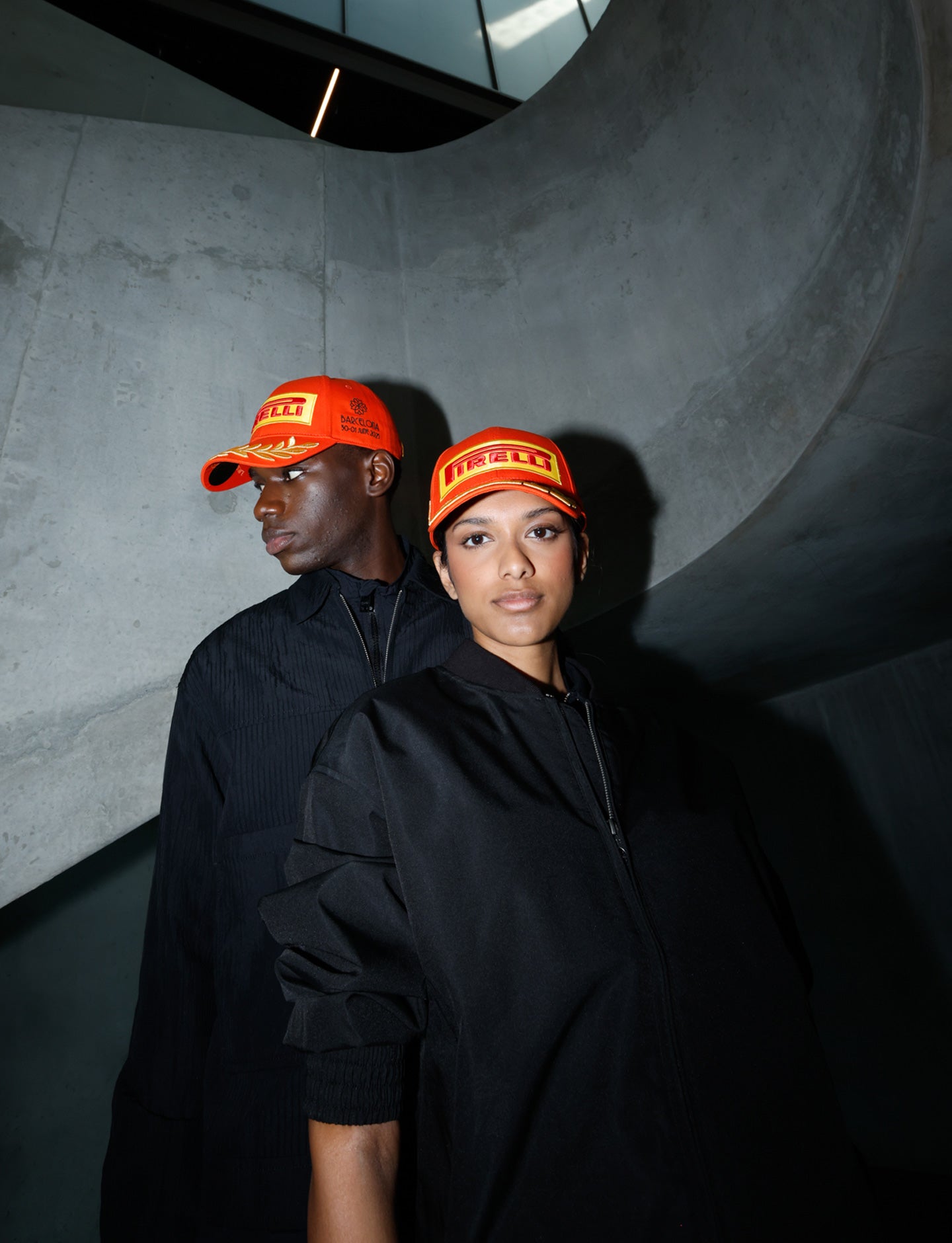 Two people wearing orange Pirelli caps and black outfits against a dark concrete wall.