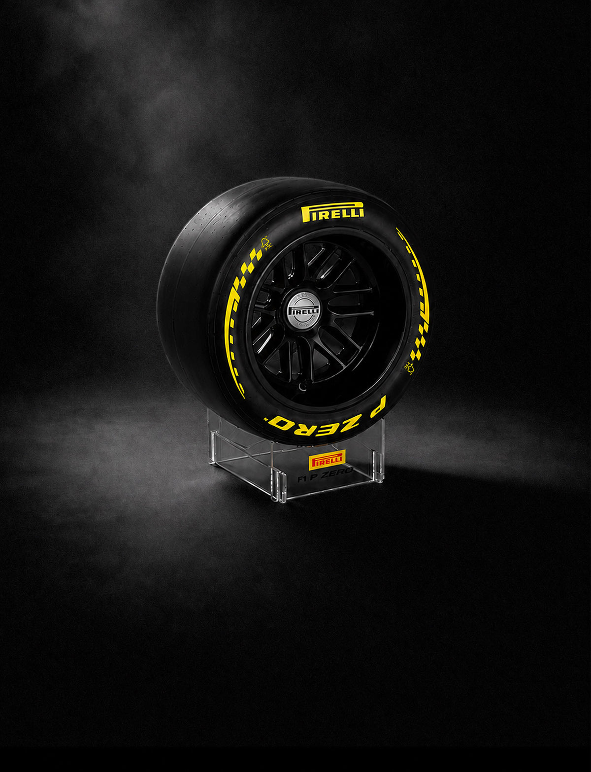 A Pirelli P Zero F1® race tire displayed upright on a clear acrylic stand against a dark, smoky studio background. The tire is black with bright yellow lettering and checkered accents, and the Pirelli logo is visible on both the tire sidewall and the center cap.